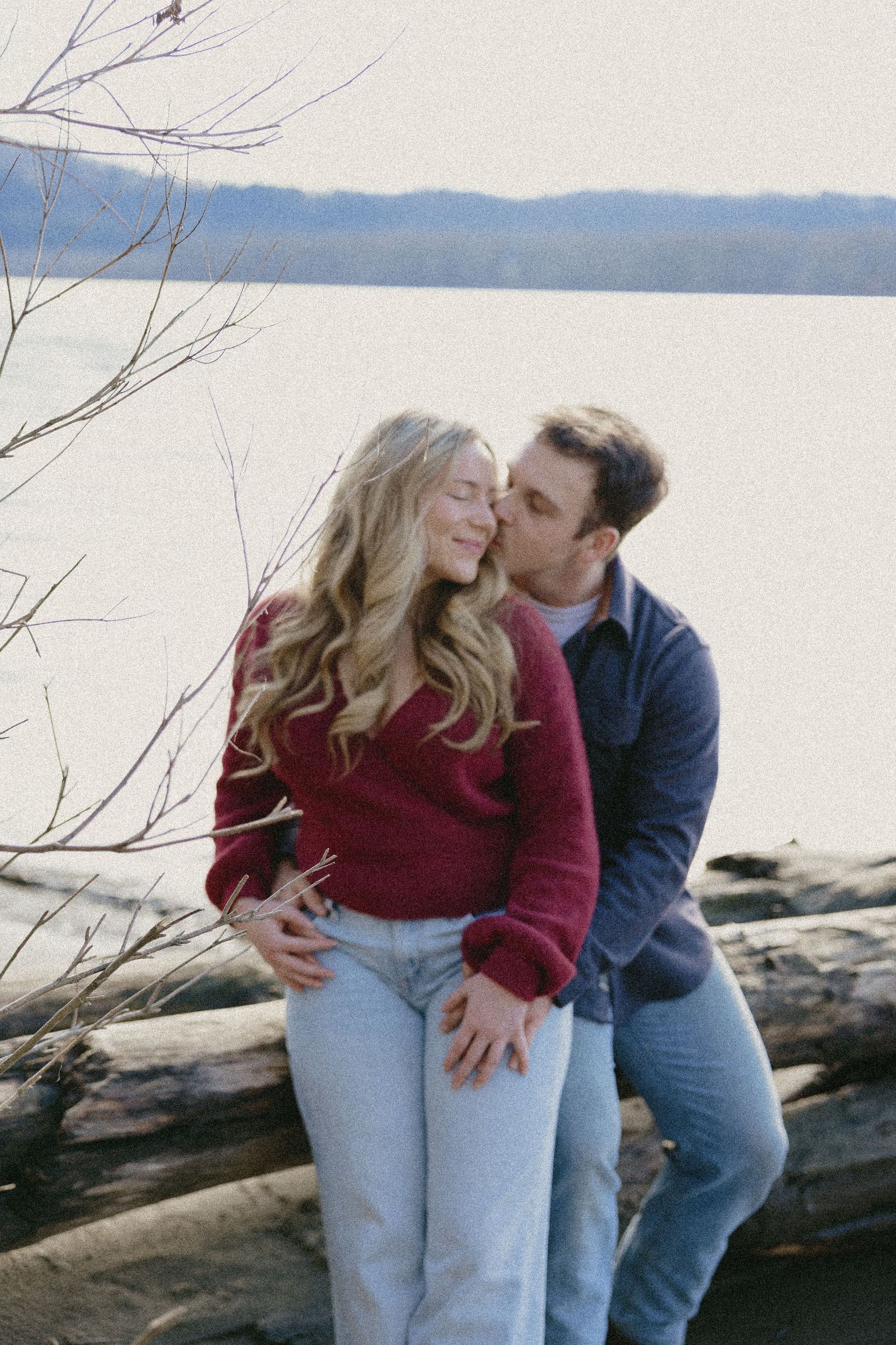 Man kissing woman on cheek while sitting on driftwood logs near a serene lake in winter.