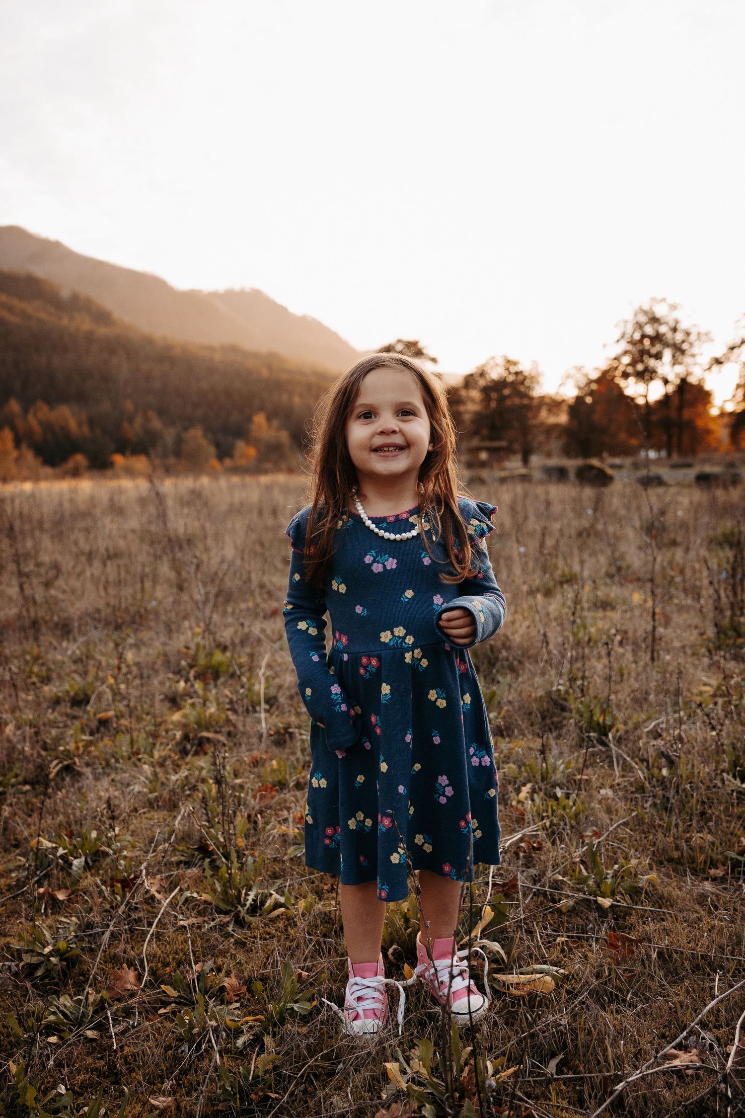 A cheerful young girl in a navy floral dress stands smiling in a golden autumn meadow.