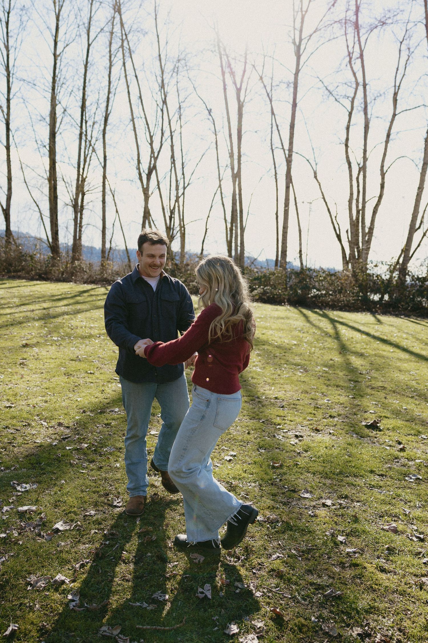 Couple spinning and laughing together on a bright grassy field with bare winter trees casting shadows.