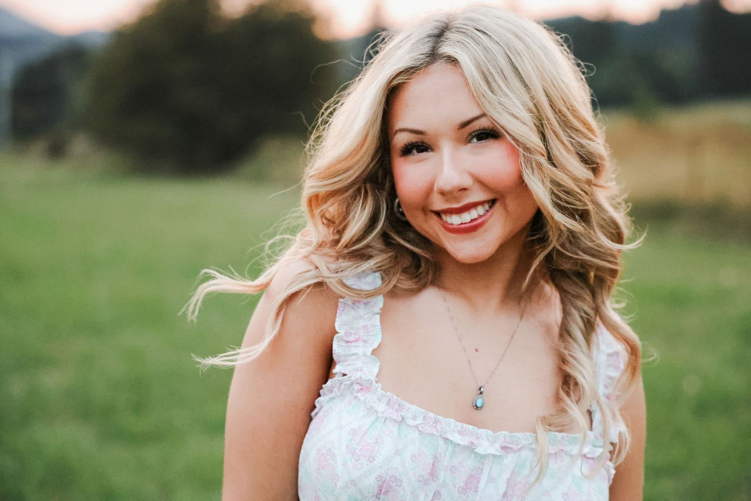 Smiling blonde woman in white floral dress outdoors in green field with mountains in background.