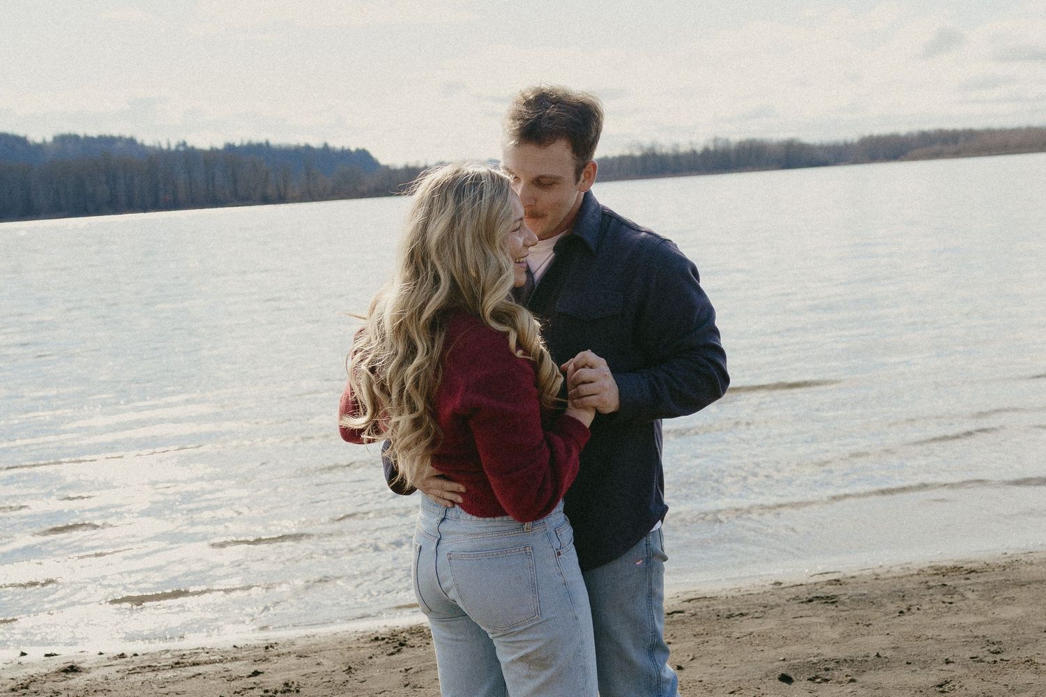Couple standing close together holding hands on a sandy lake shore in casual fall clothing.
