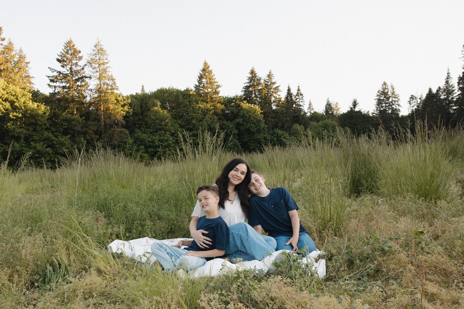 A woman and two children sit on a blanket in a sunlit meadow with tall grass and trees in the background.