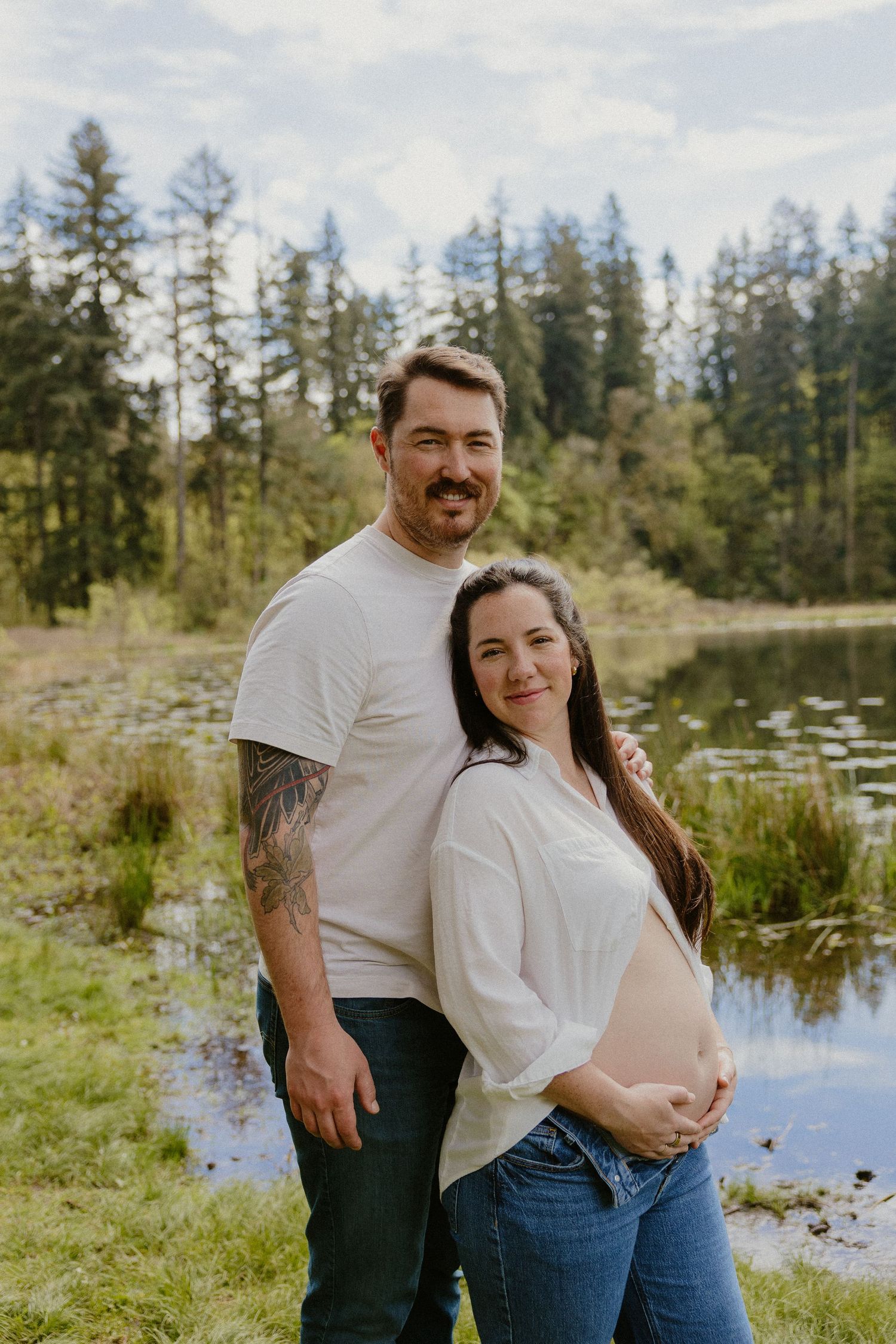 Expecting couple smiling together by a peaceful forest lake.
