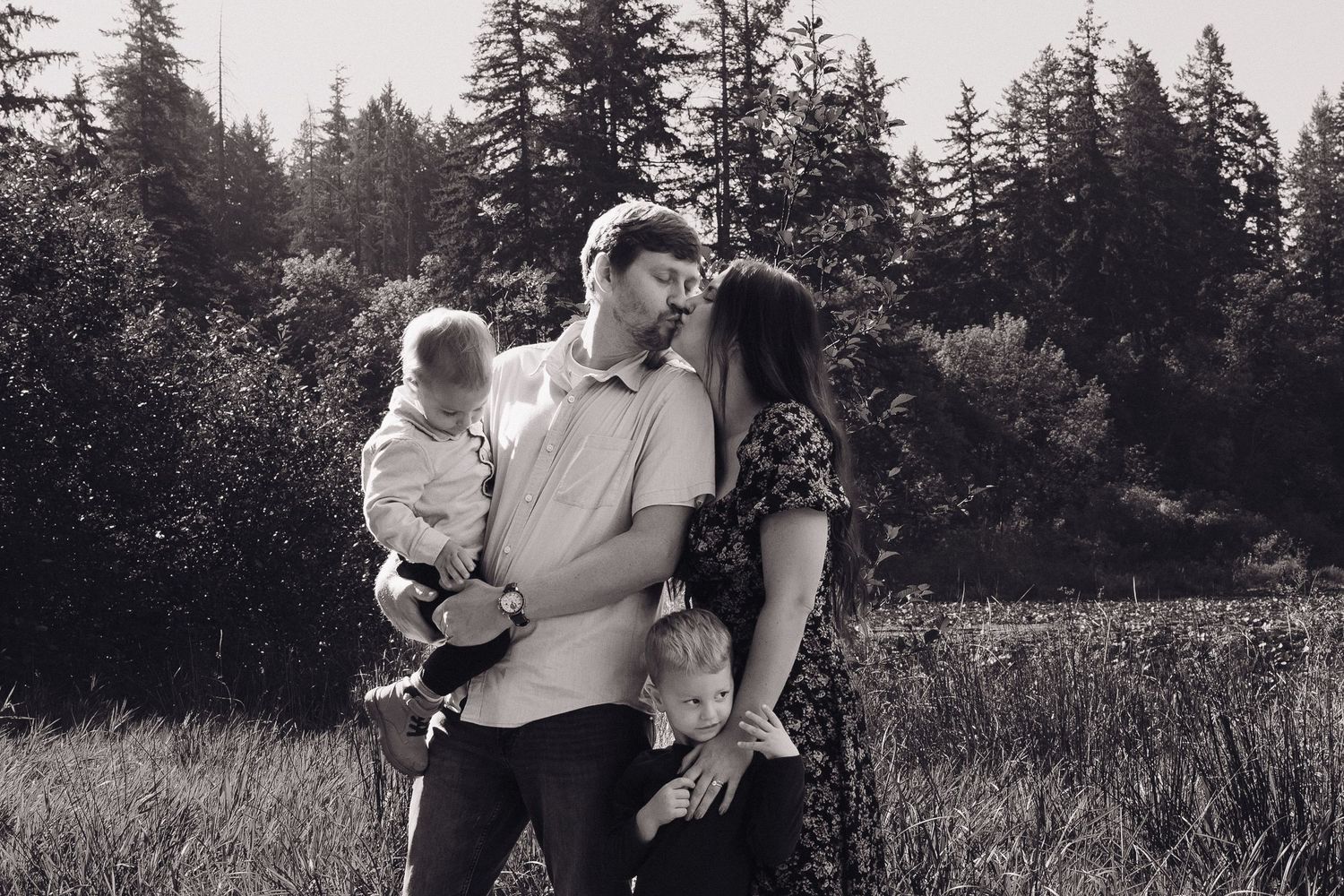Black and white photo of couple kissing while holding two young children in open field.
