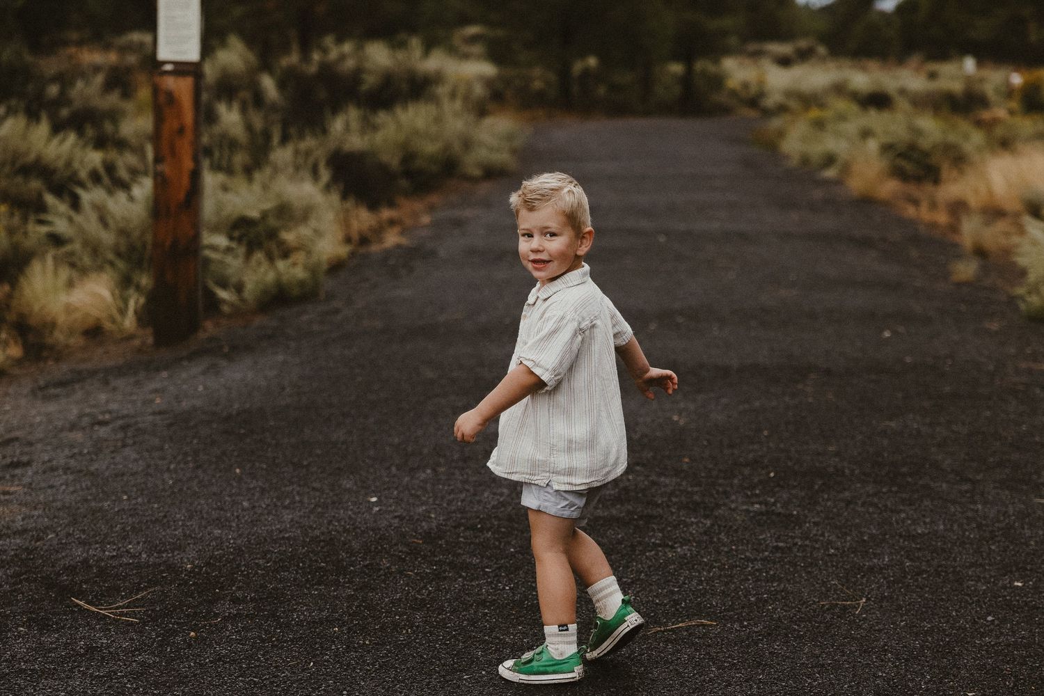 Cute toddler boy in white shirt and green sneakers walking on rural gravel road.