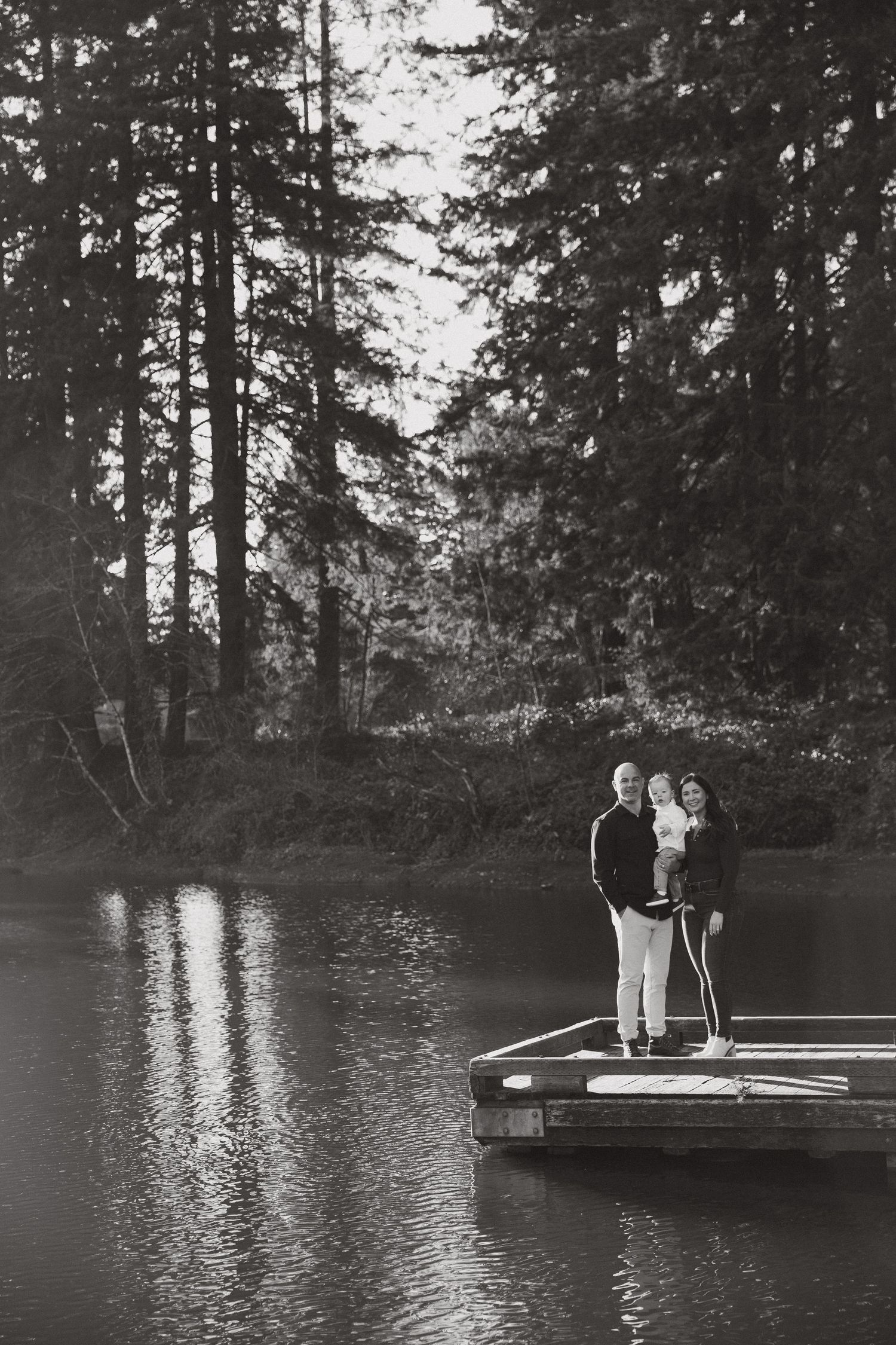 Black and white family portrait on a wooden dock overlooking a calm lake with tall trees.