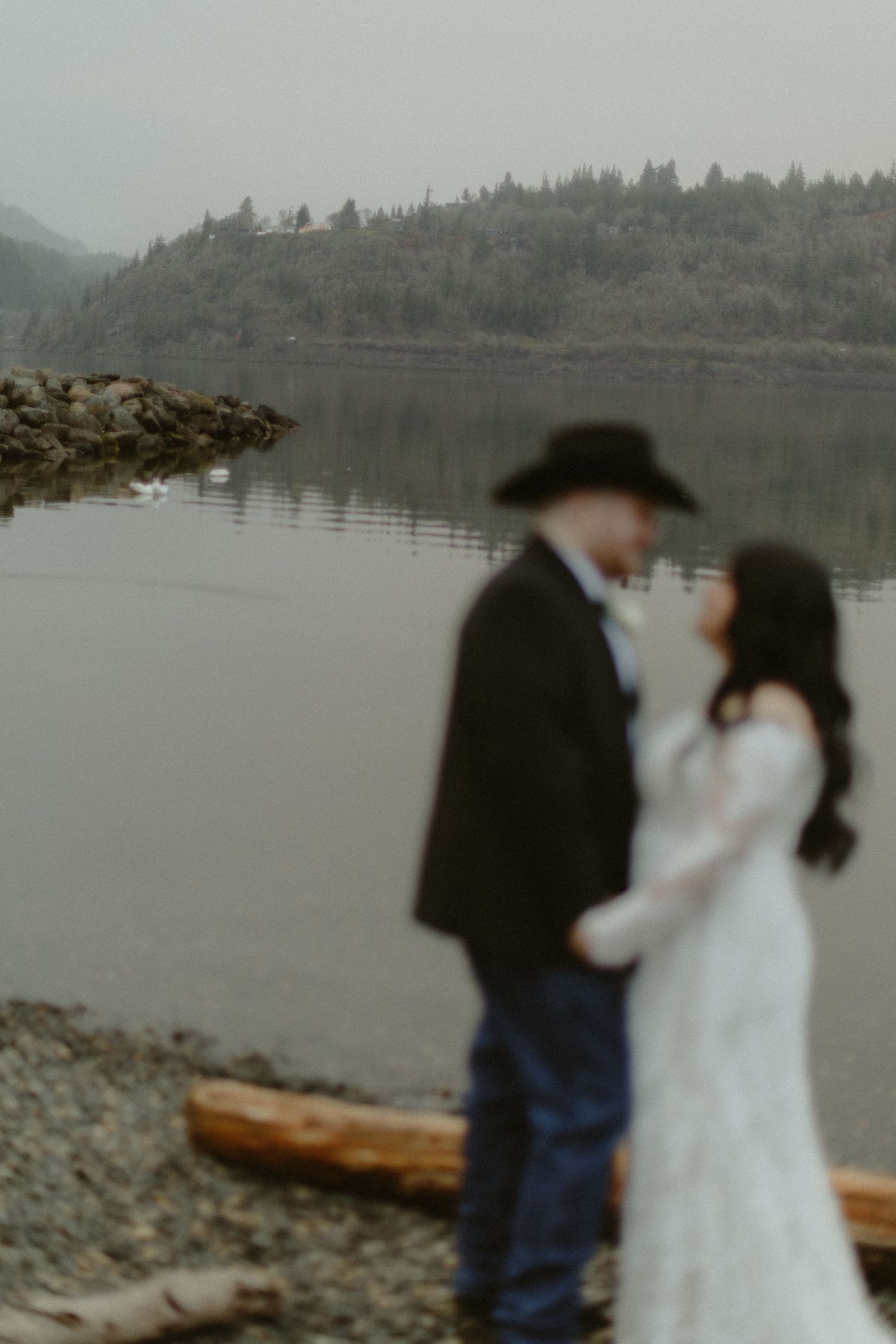 Artistic blurred foreground of wedding couple standing close on a rocky shore with calm lake backdrop.