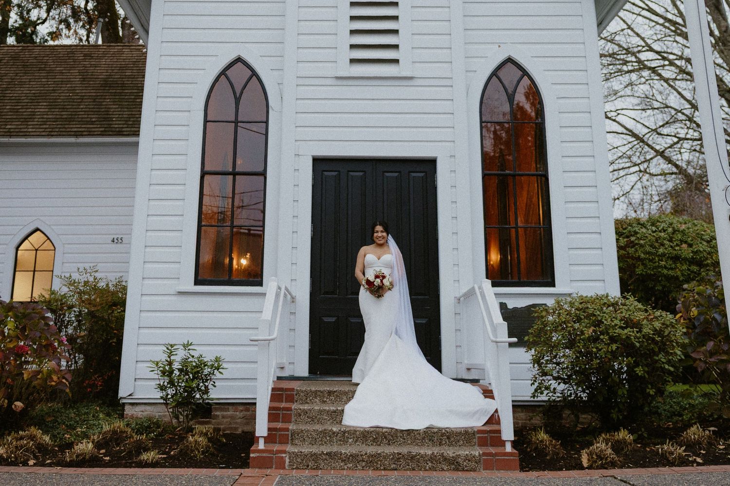 Bride in white mermaid gown with veil poses on steps of white chapel with black doors.