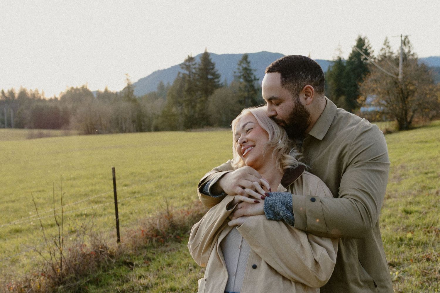 Man embracing woman from behind in open green field with mountains in background.