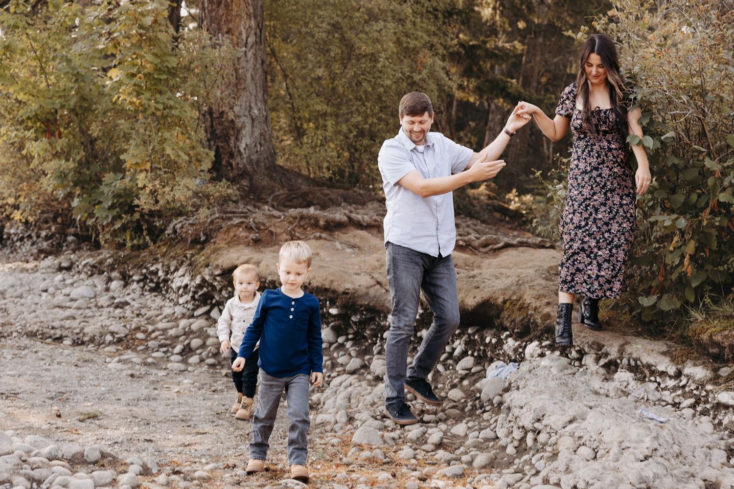 Family of four walking on rocky riverbank through colorful autumn forest, parents holding hands.