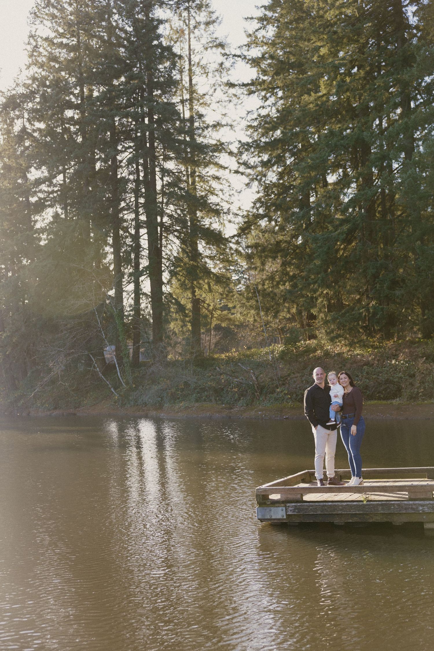 Family of three standing together on a wooden lake dock surrounded by tall evergreen trees in golden light.