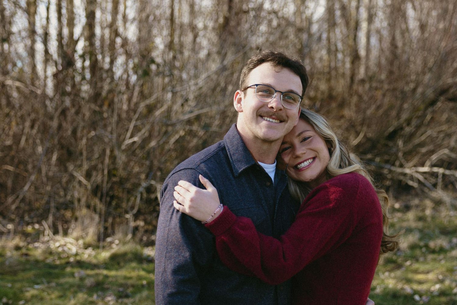 Happy couple hugging and laughing together outdoors in front of bare winter trees and greenery.