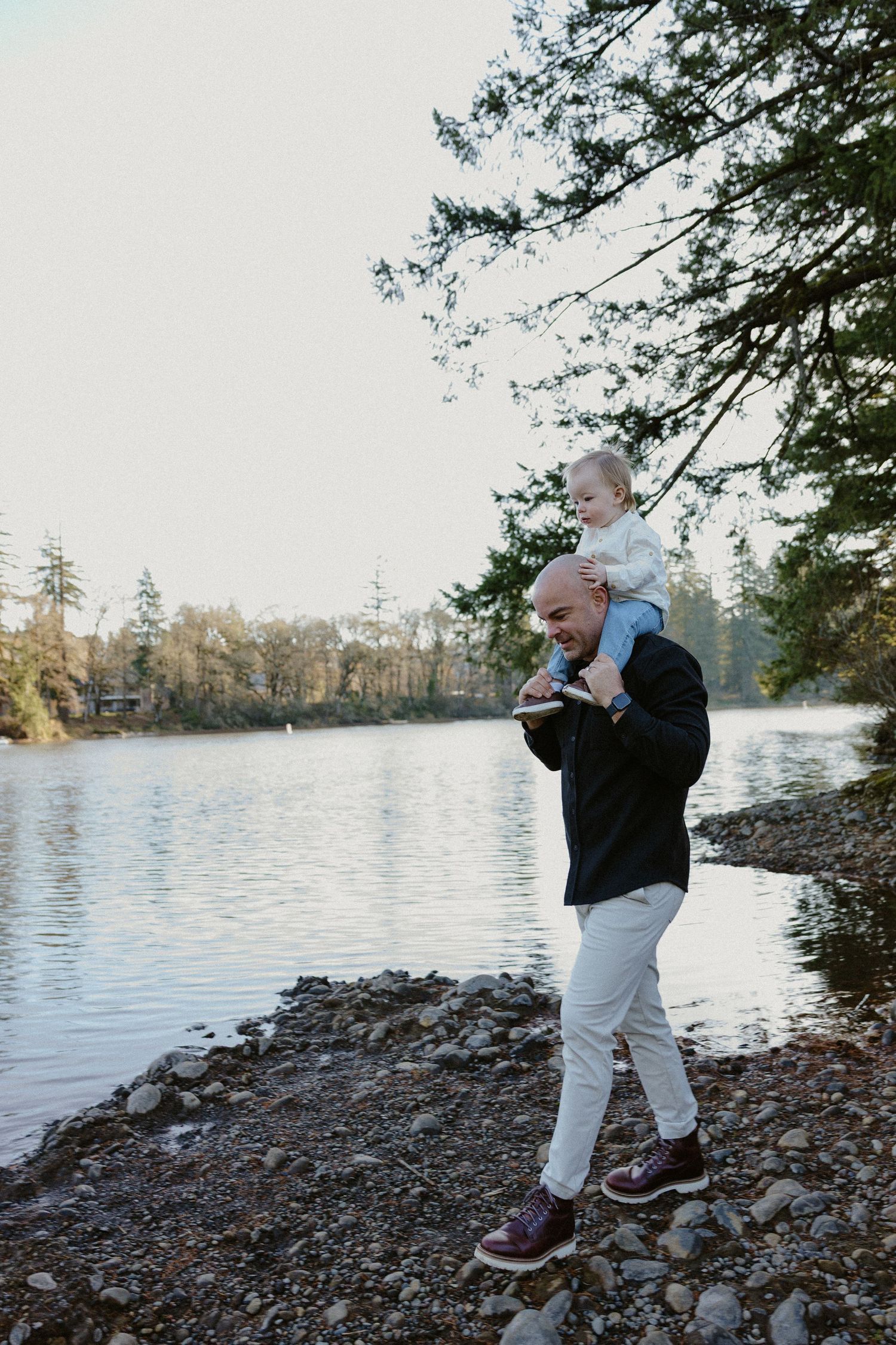 Father carrying toddler on shoulders along a rocky lakeside path beneath tall evergreen trees.