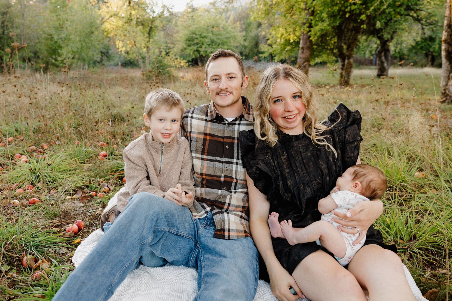 A happy family of four sits together on a blanket in an autumn orchard smiling at the camera.