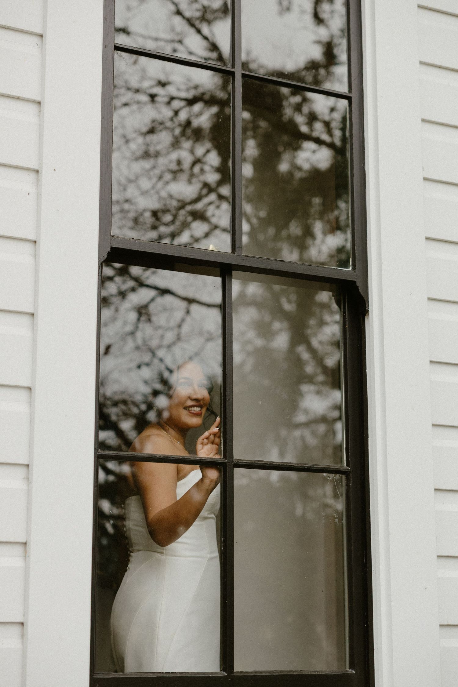 Smiling bride in white gown visible through a large black-framed window of a white clapboard building.