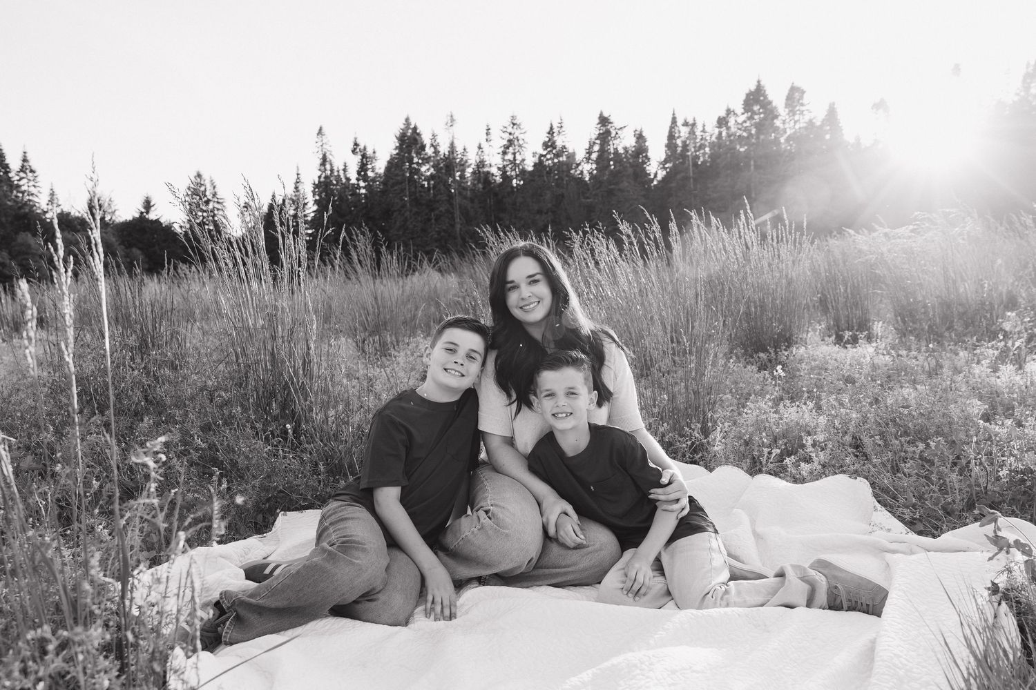 Black and white photo of a mother sitting in a field with her two children, smiling together on a white blanket.