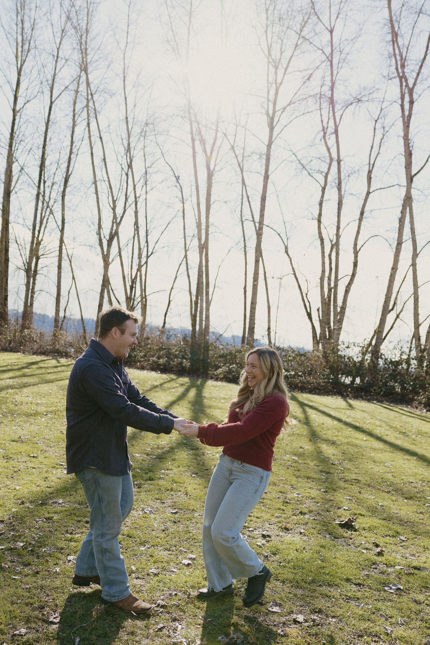 Couple holding hands and dancing playfully on sunny grass with tall bare trees behind them.