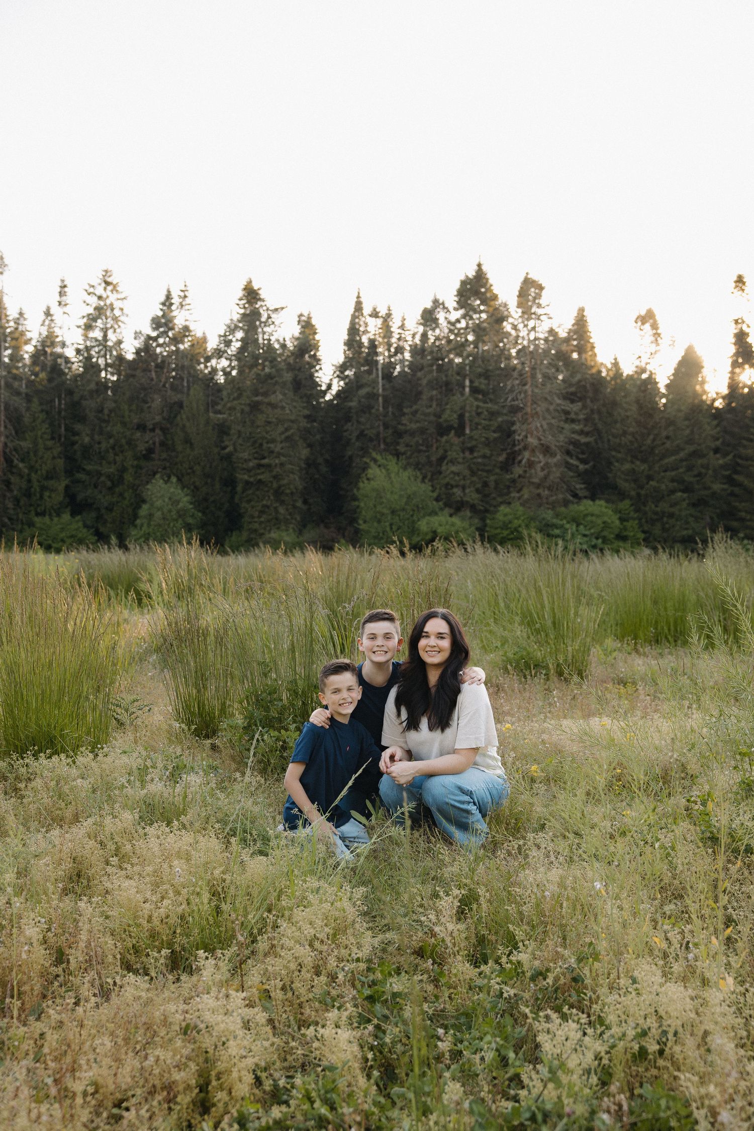 Family of three posing in a golden meadow with tall grass and pine trees at sunset.
