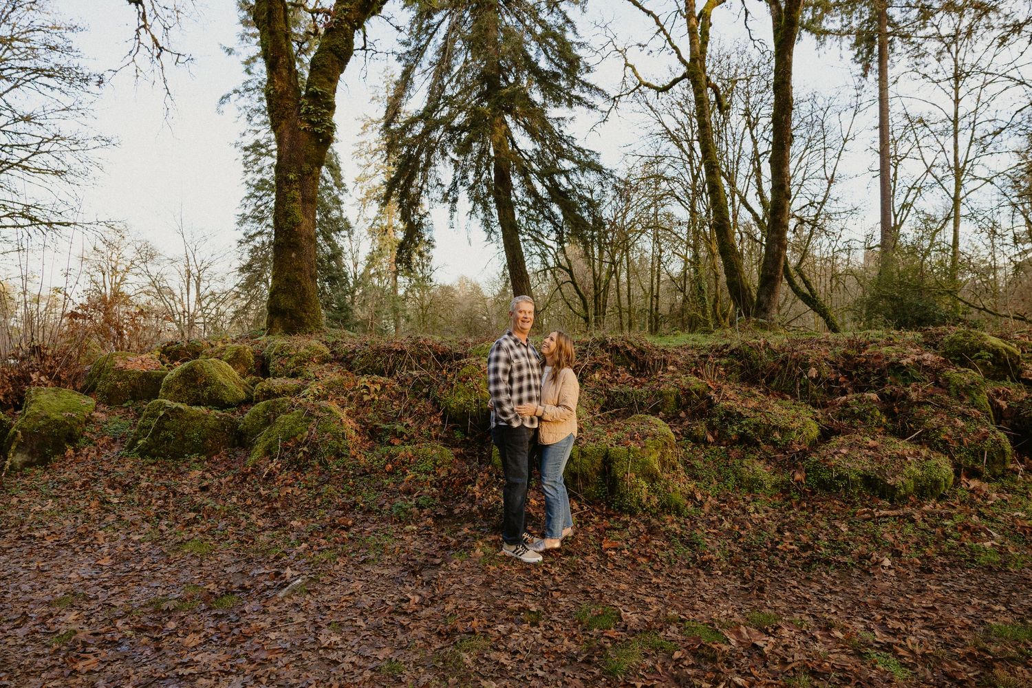 Smiling couple embracing on moss-covered rocks in a lush winter woodland forest.