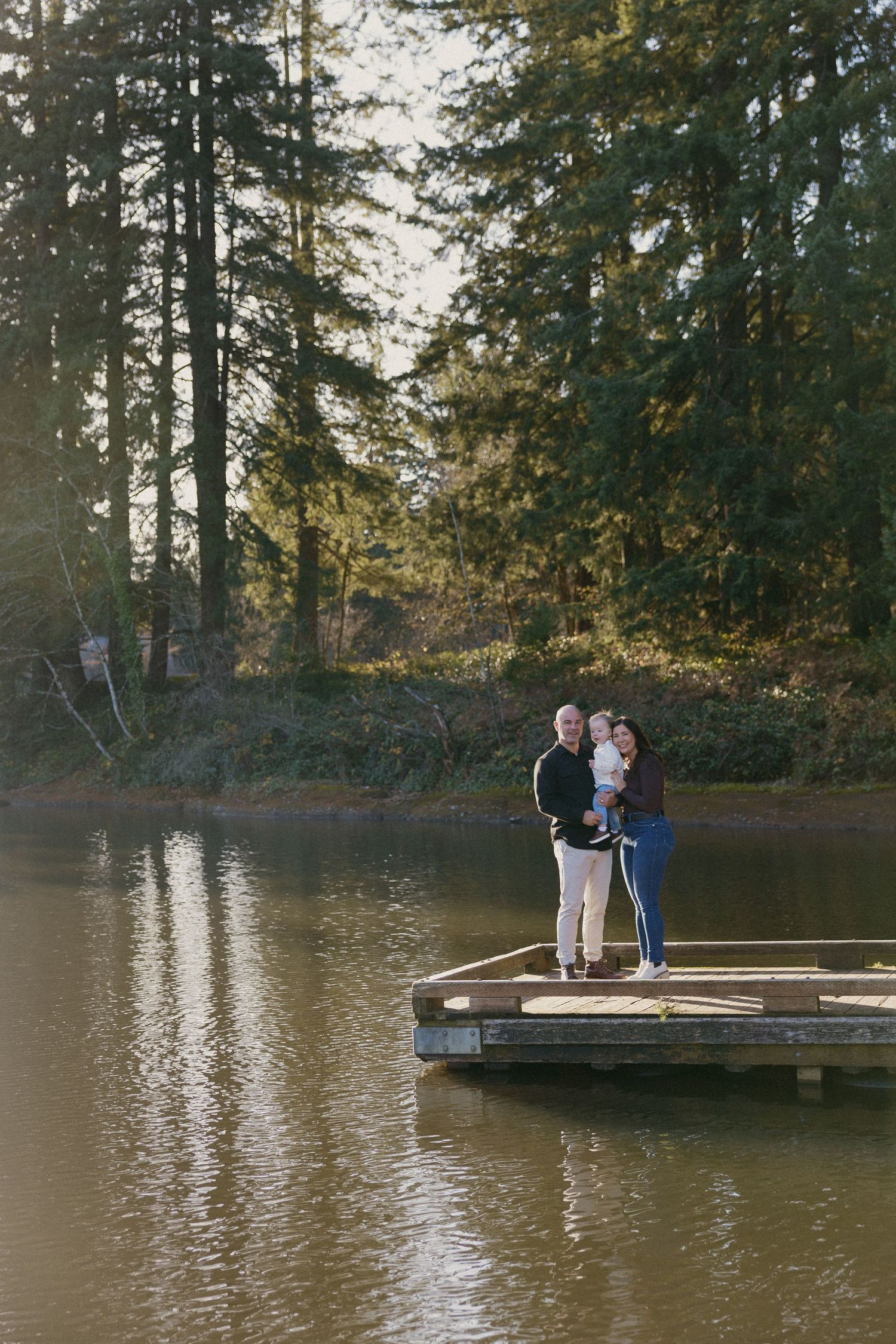 Family of three stands on a wooden dock surrounded by a serene lake and tall evergreen trees.