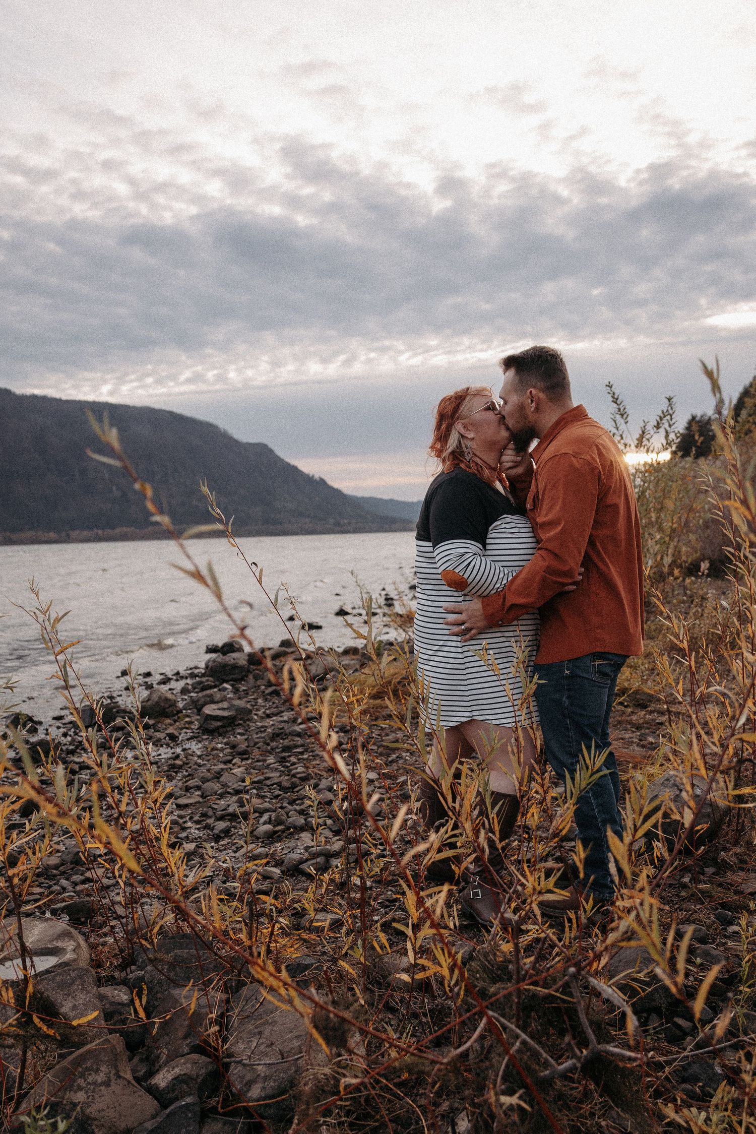 Couple embracing and kissing near a rocky riverbank with dramatic cloudy sky.