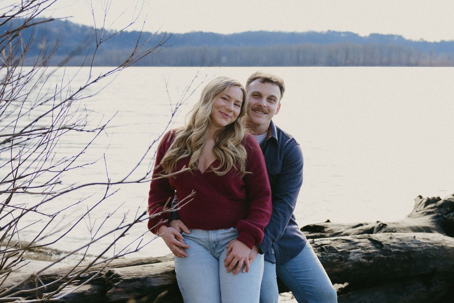 Smiling couple sitting on driftwood by a calm lake, holding hands during fall engagement session.