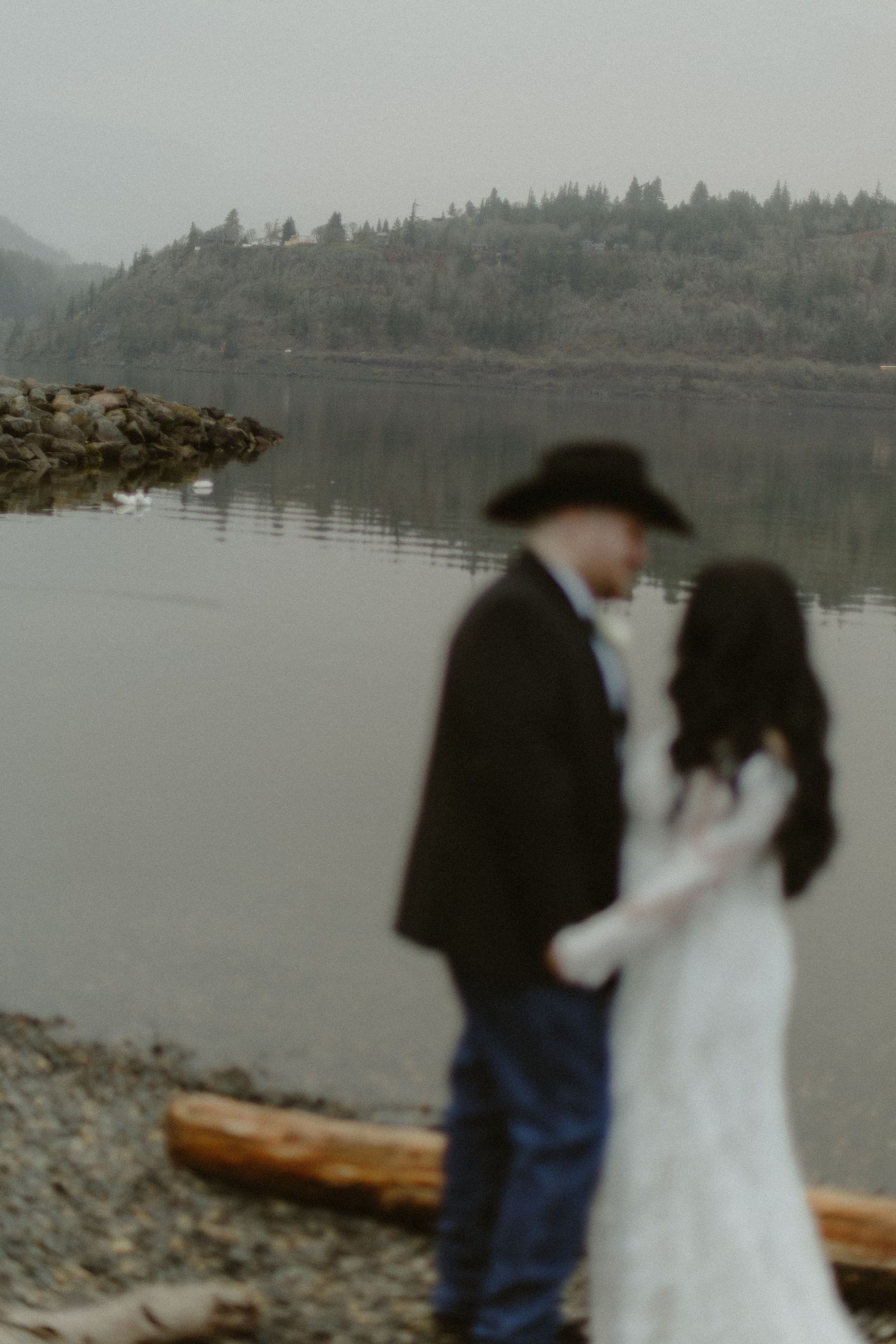 Defocused wedding couple holding hands by a calm lake, groom wearing cowboy hat and dark suit.