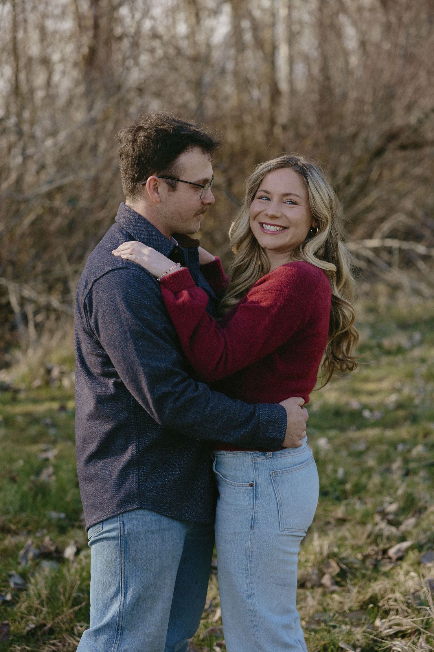 Couple embracing and smiling in a natural outdoor setting with bare trees in background.