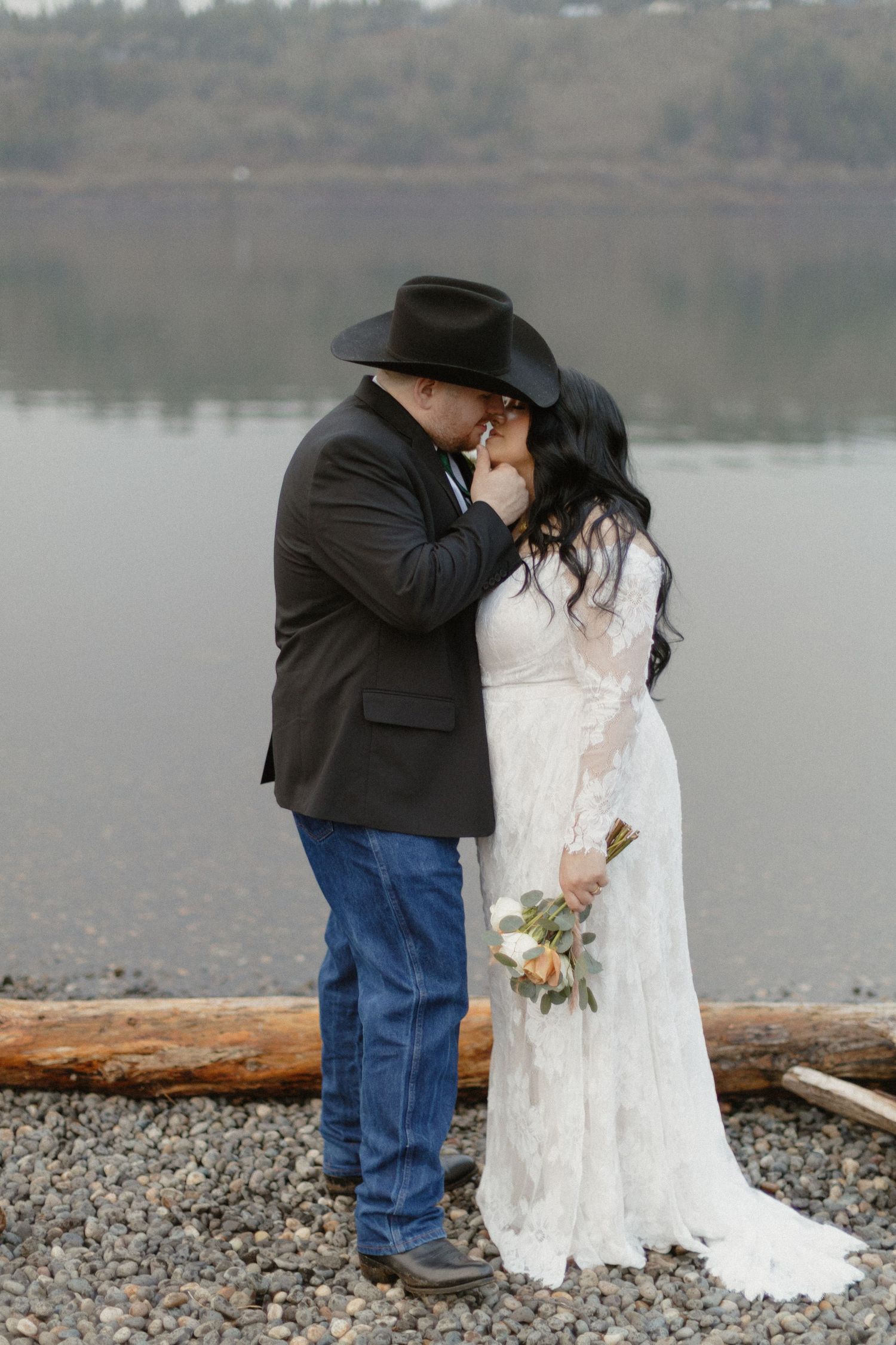 Bride and groom share a kiss by a misty lake, she in lace gown holding bouquet, he in black jacket and cowboy hat.