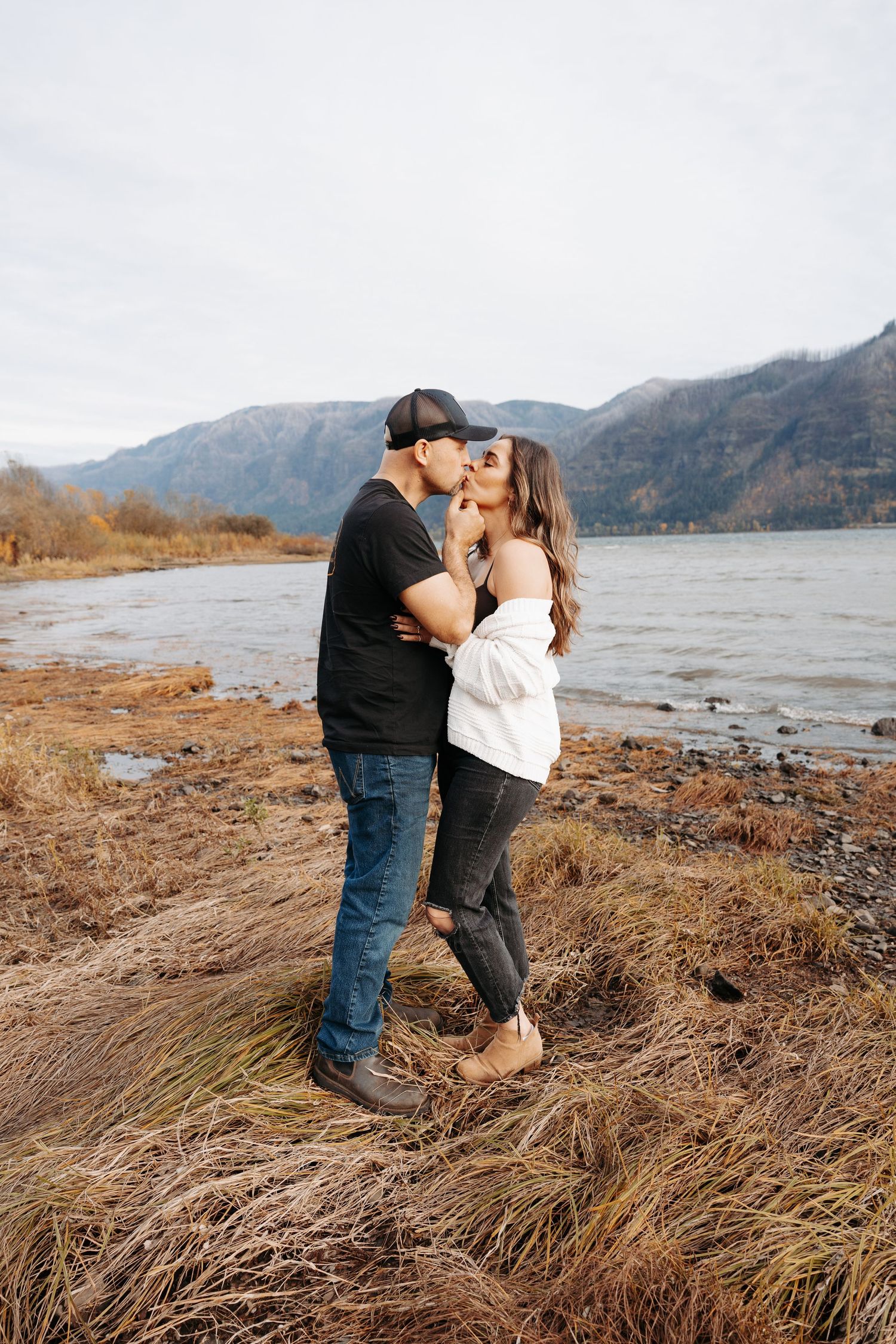 Couple sharing a romantic kiss by a river with mountain backdrop in autumn.