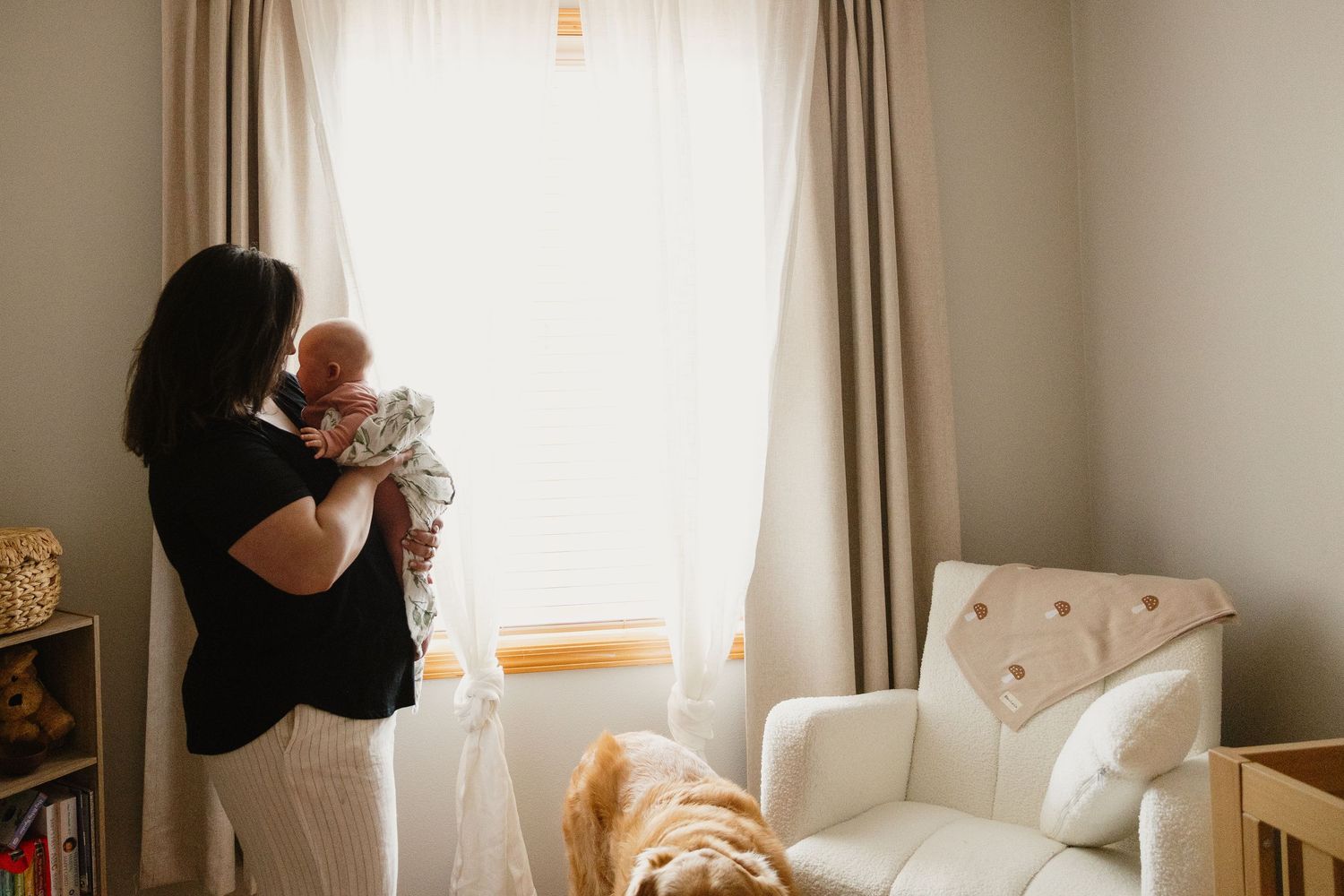 Mother standing by nursery window holding newborn baby with golden retriever nearby.