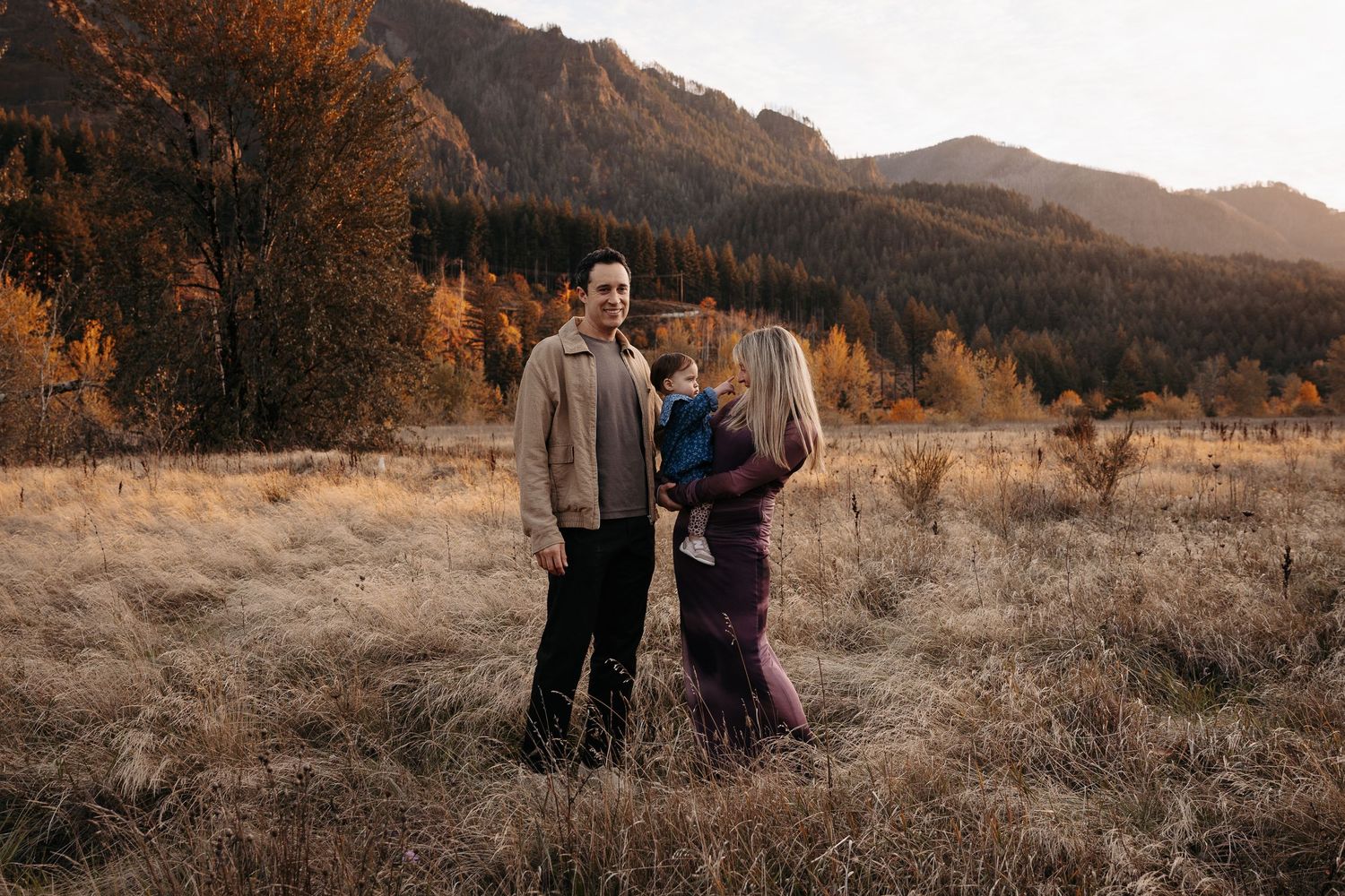 A couple holding their baby stands together in a golden autumn field with mountain views.