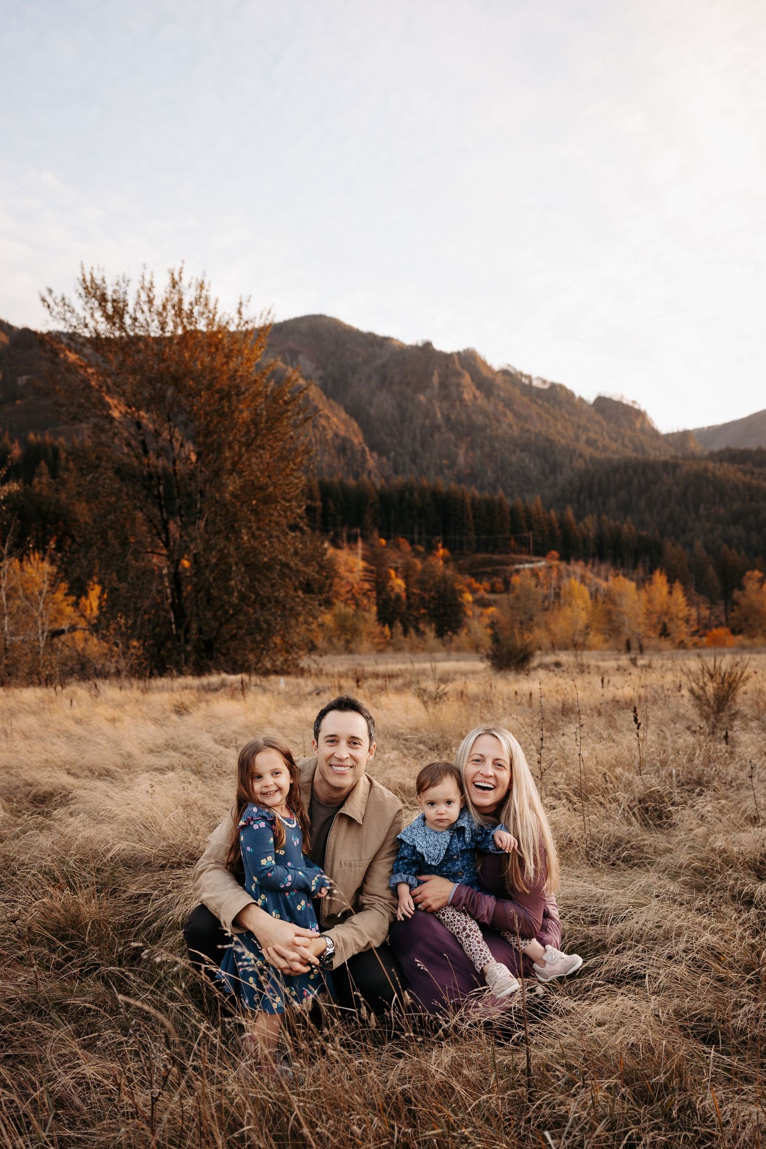 Happy family of four sitting in dry grass field with colorful autumn mountain landscape behind them.
