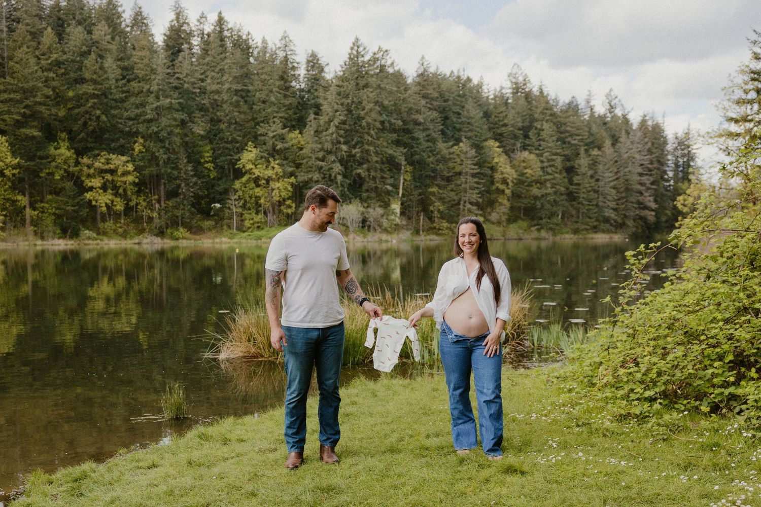Expecting couple walking by a forest pond holding tiny baby clothes during maternity shoot.
