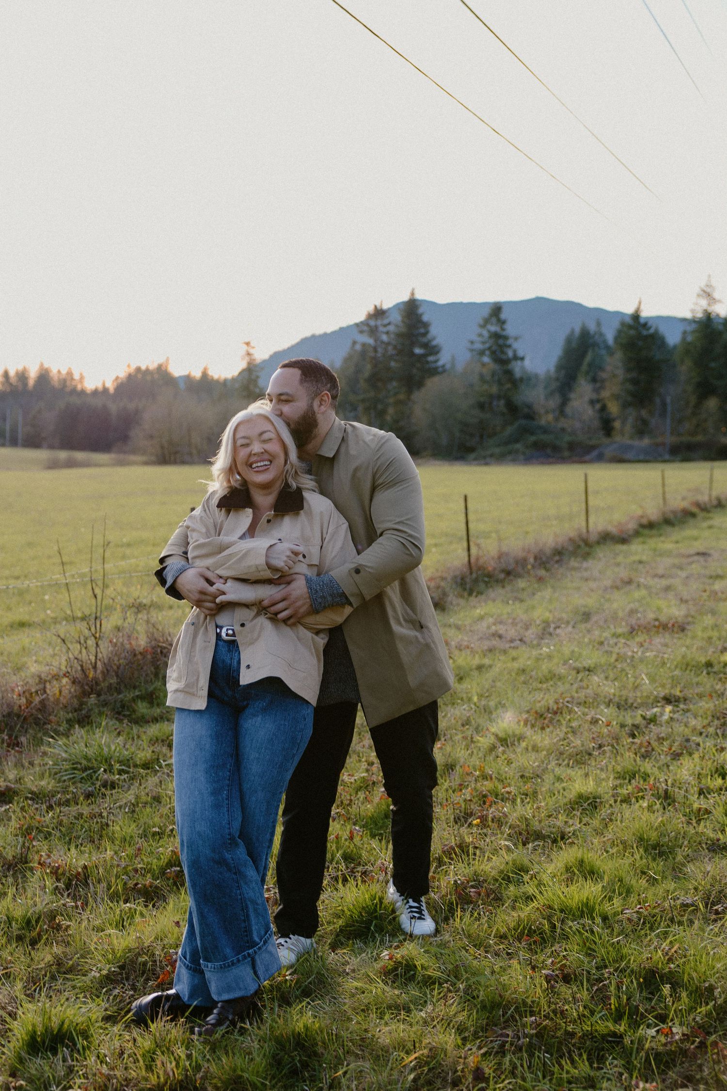 Couple laughing and embracing in a green meadow at golden hour sunset.