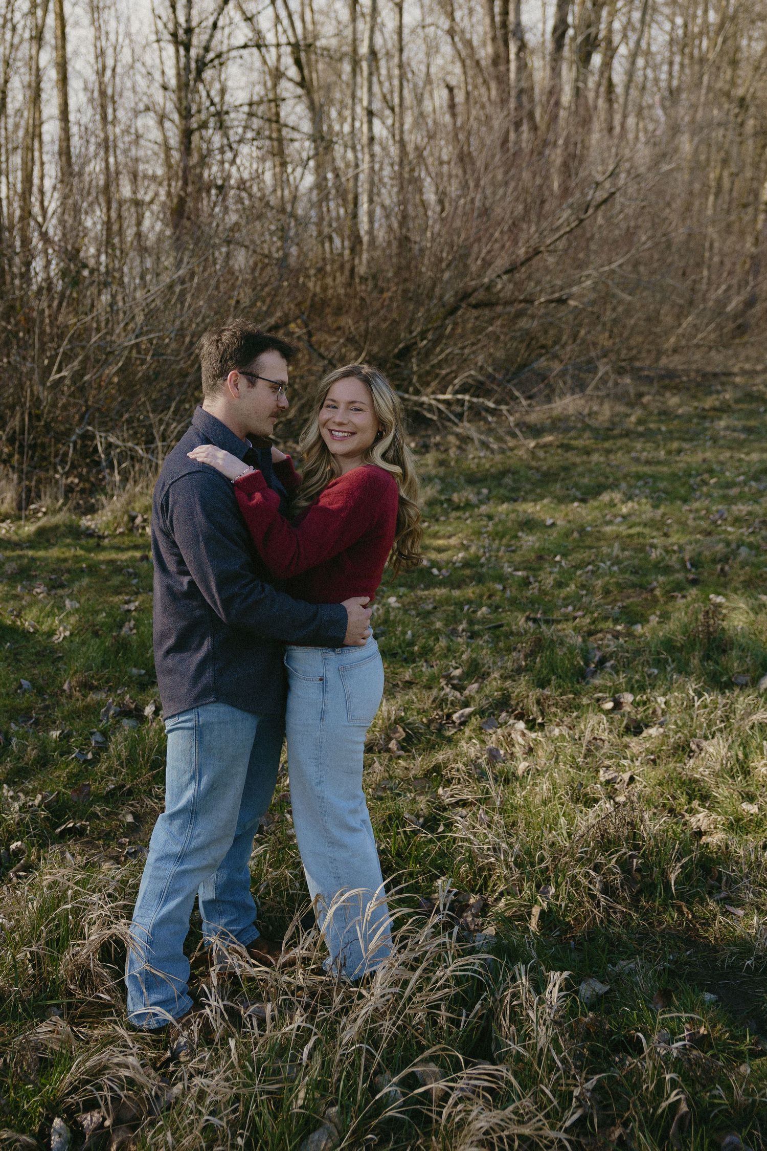 Happy couple embracing in a sunlit field with bare winter trees, woman in red sweater and jeans.