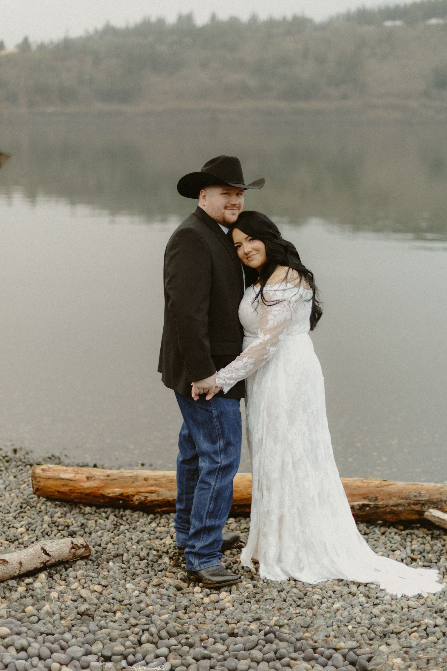 A groom in a black cowboy hat and bride in lace gown pose by a rocky lakeside.