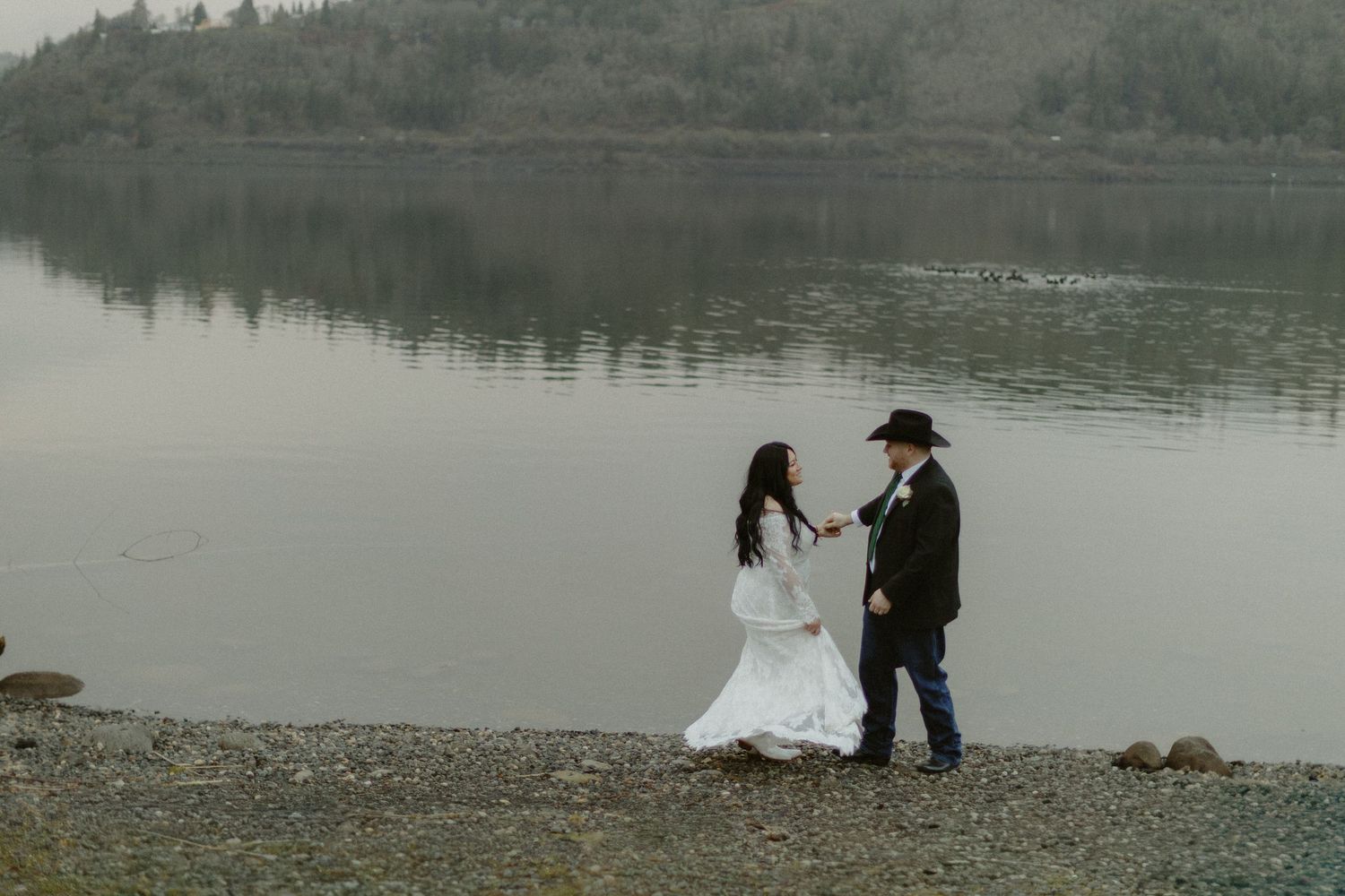 Wedding couple facing each other on a rocky beach beside a serene misty lake holding hands.