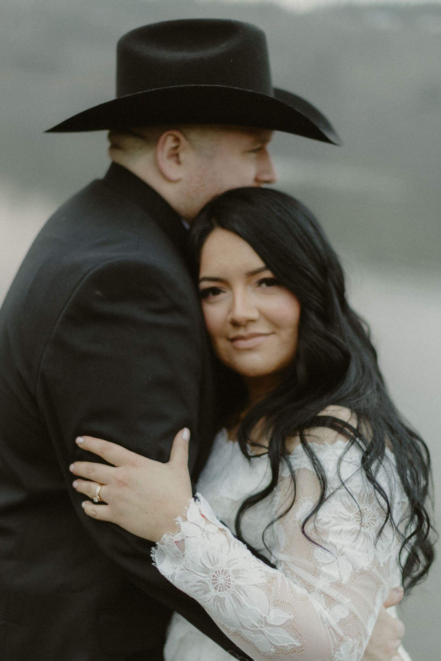 Bride in white lace gown smiling as groom in black cowboy hat embraces her during wedding portraits.