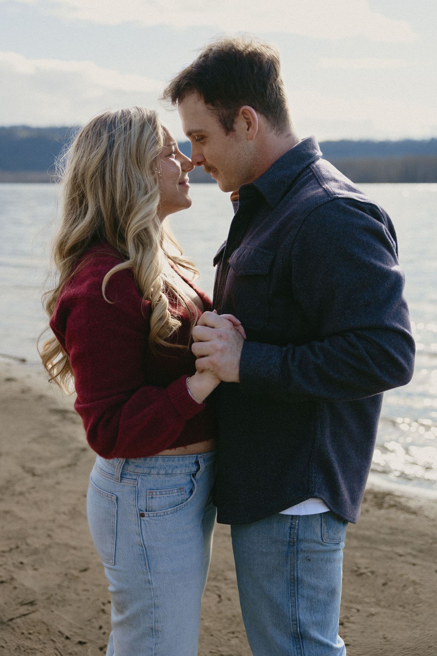 Romantic couple holding hands and leaning close on a sandy lakeside beach in casual fall attire.