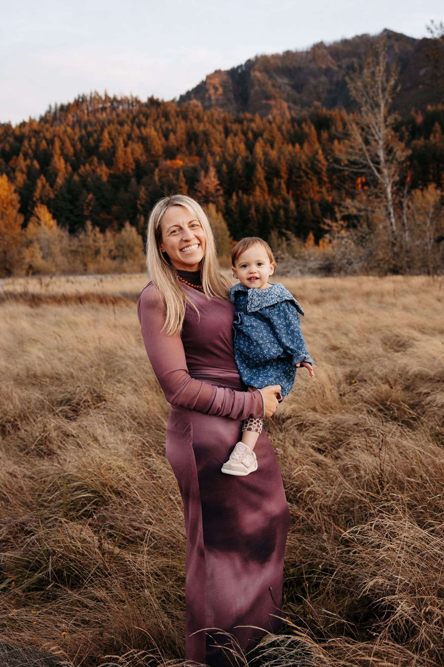 A smiling blonde mother in a mauve dress holds her baby girl in an autumn field.