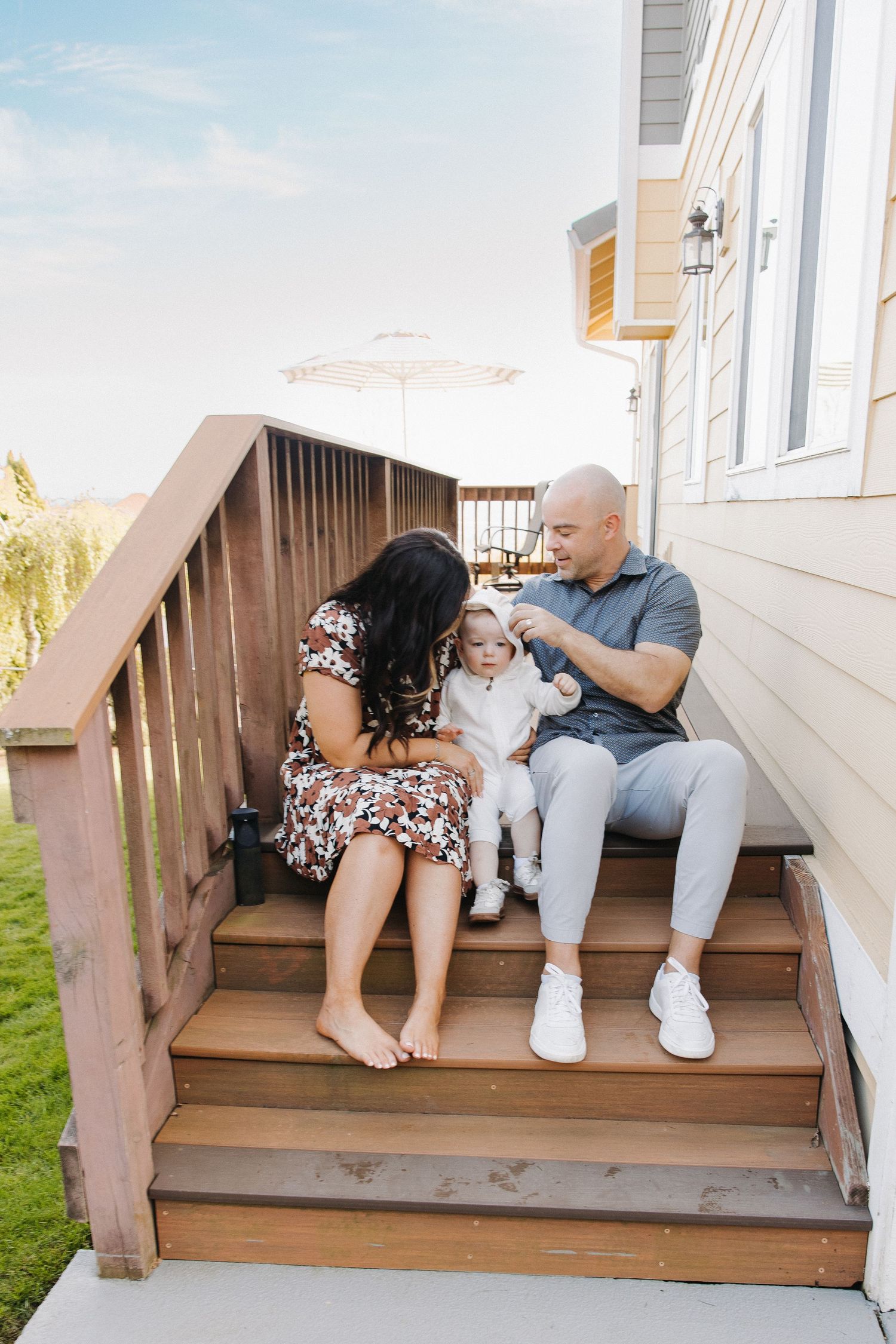 Young family with baby sitting on wooden deck steps outside their home on sunny day.