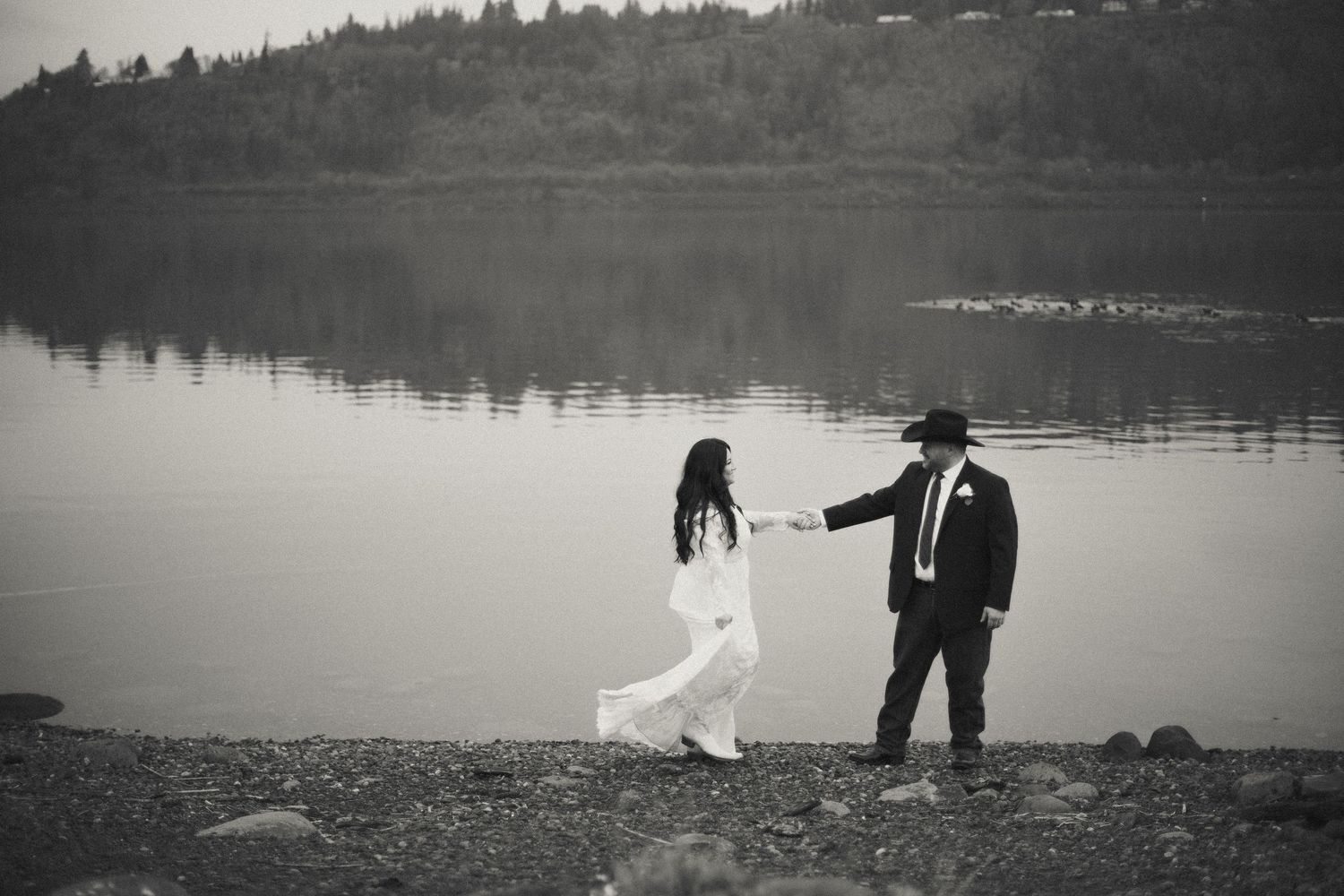 Black and white photo of bride and groom holding hands by a calm lake, groom in cowboy hat leading the way.