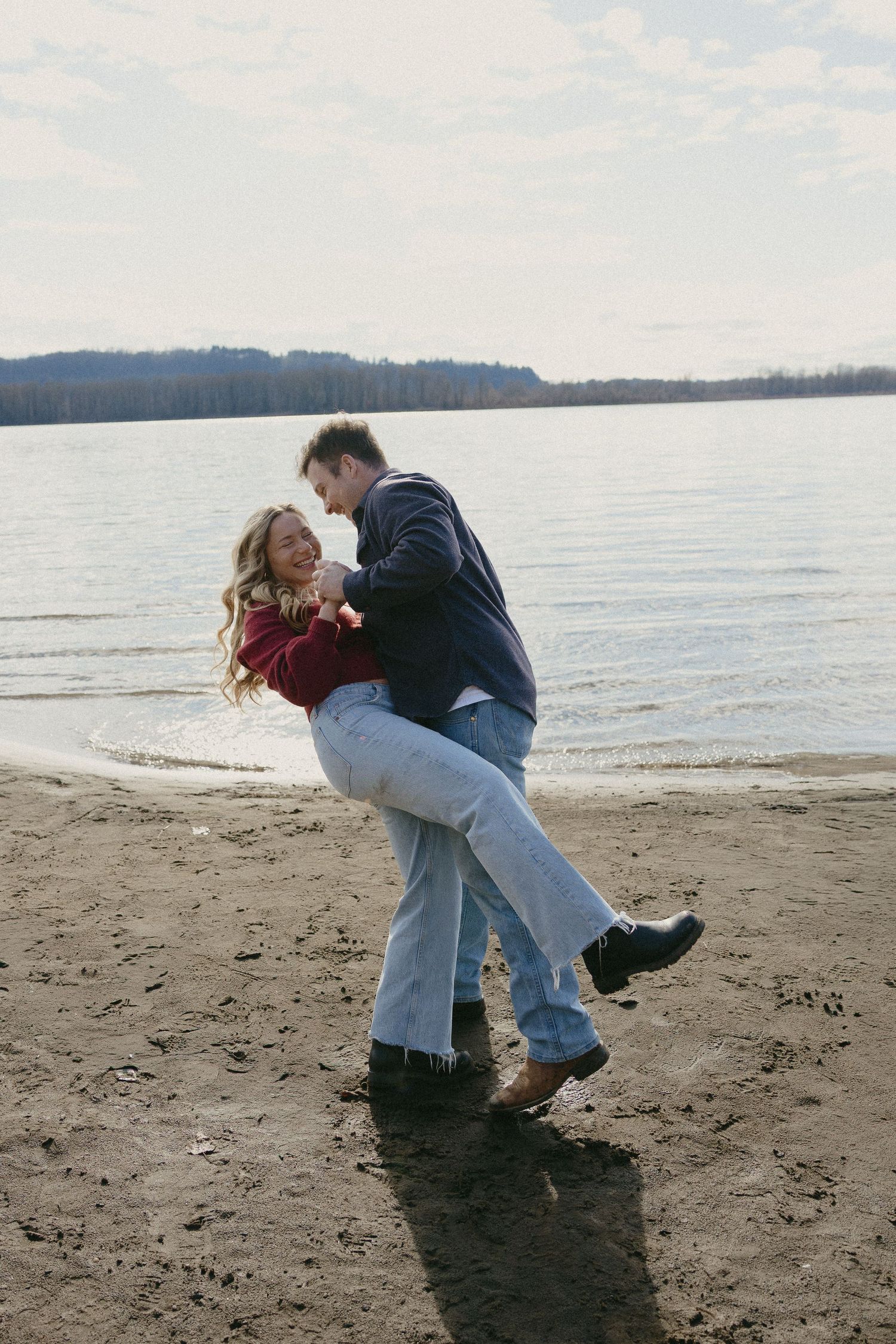 Man playfully dipping woman on a sandy lake beach, both laughing in casual jeans and sweaters.