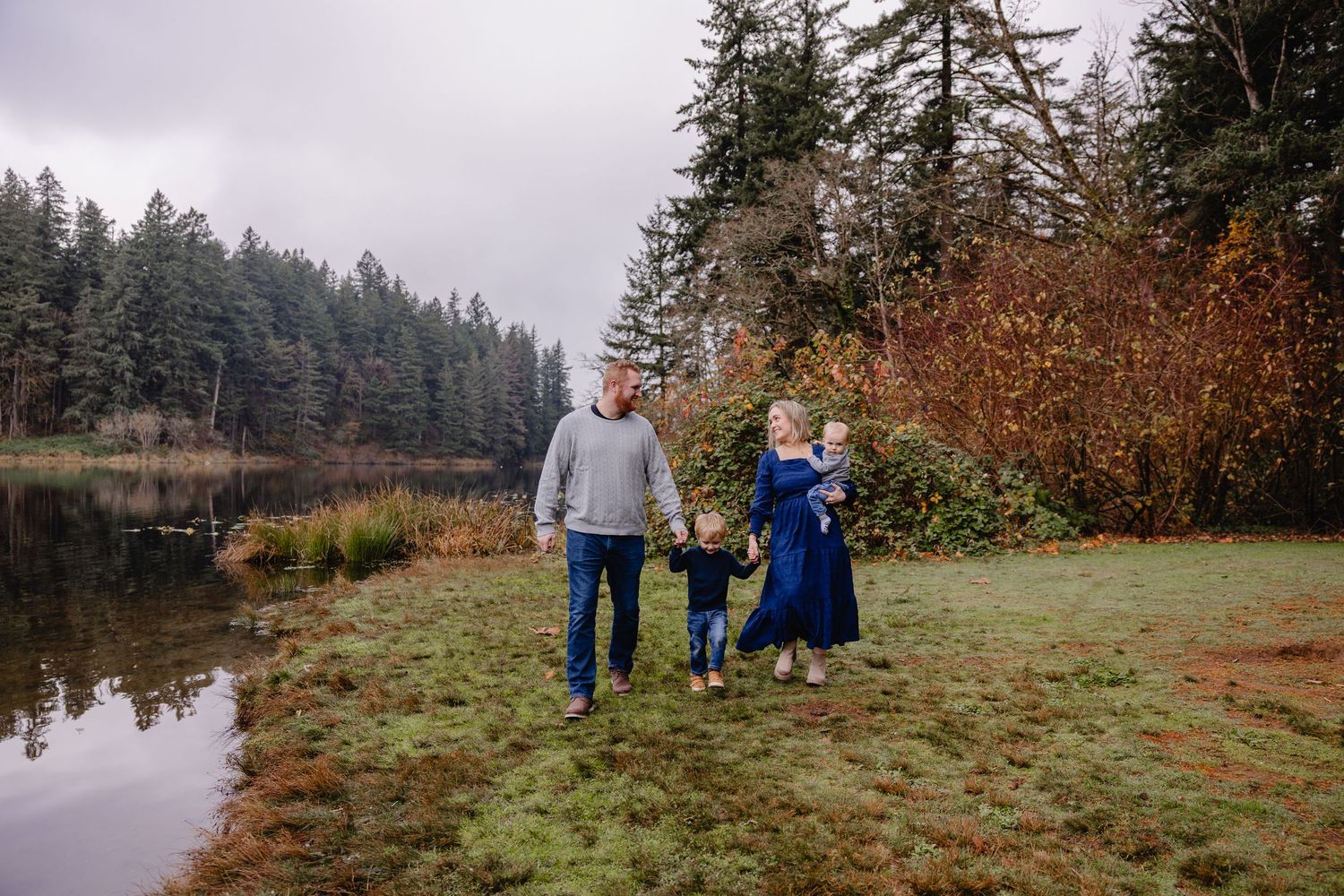 Family of three walking hand-in-hand along a misty lakeside meadow surrounded by evergreen and autumn foliage.