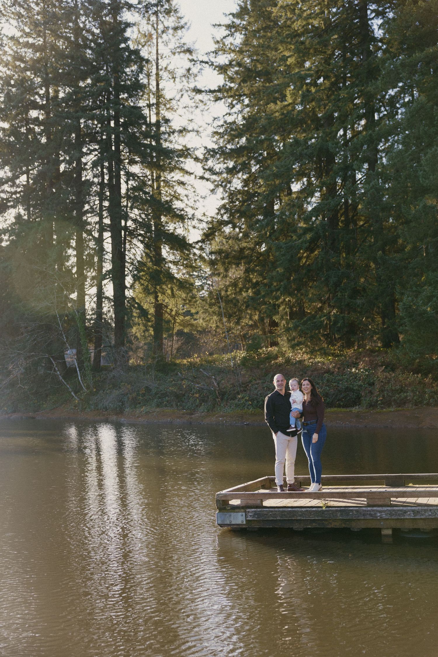 Family with baby standing on a wooden dock over a calm lake surrounded by tall evergreen trees.