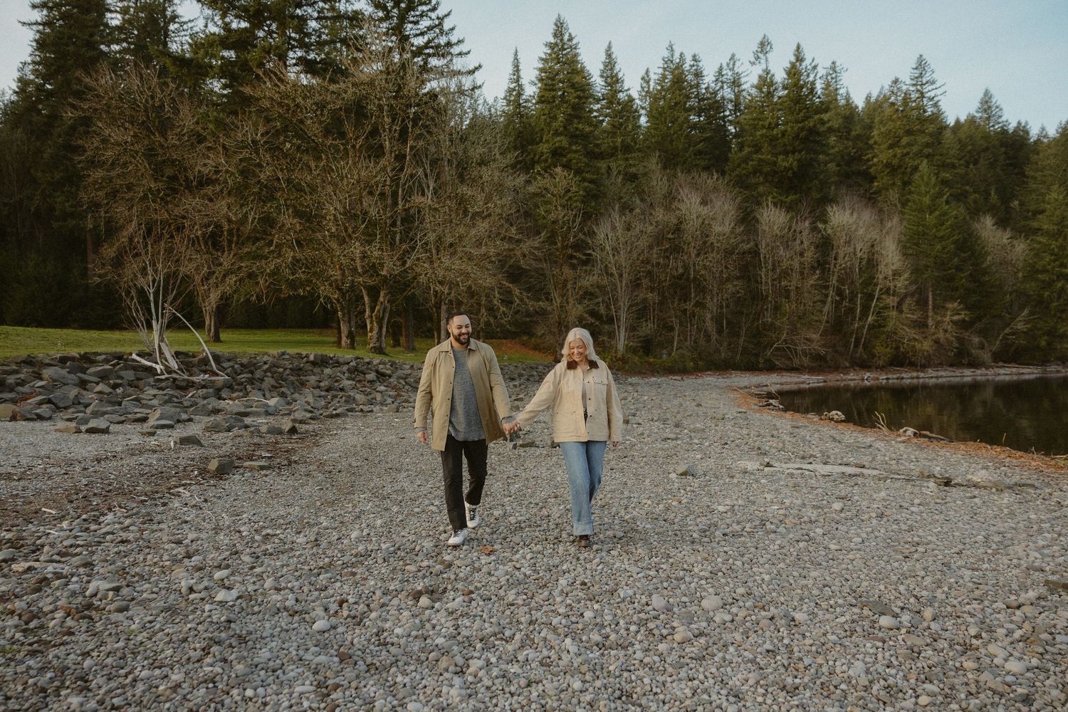 Couple holding hands while walking on a rocky beach surrounded by evergreen trees.