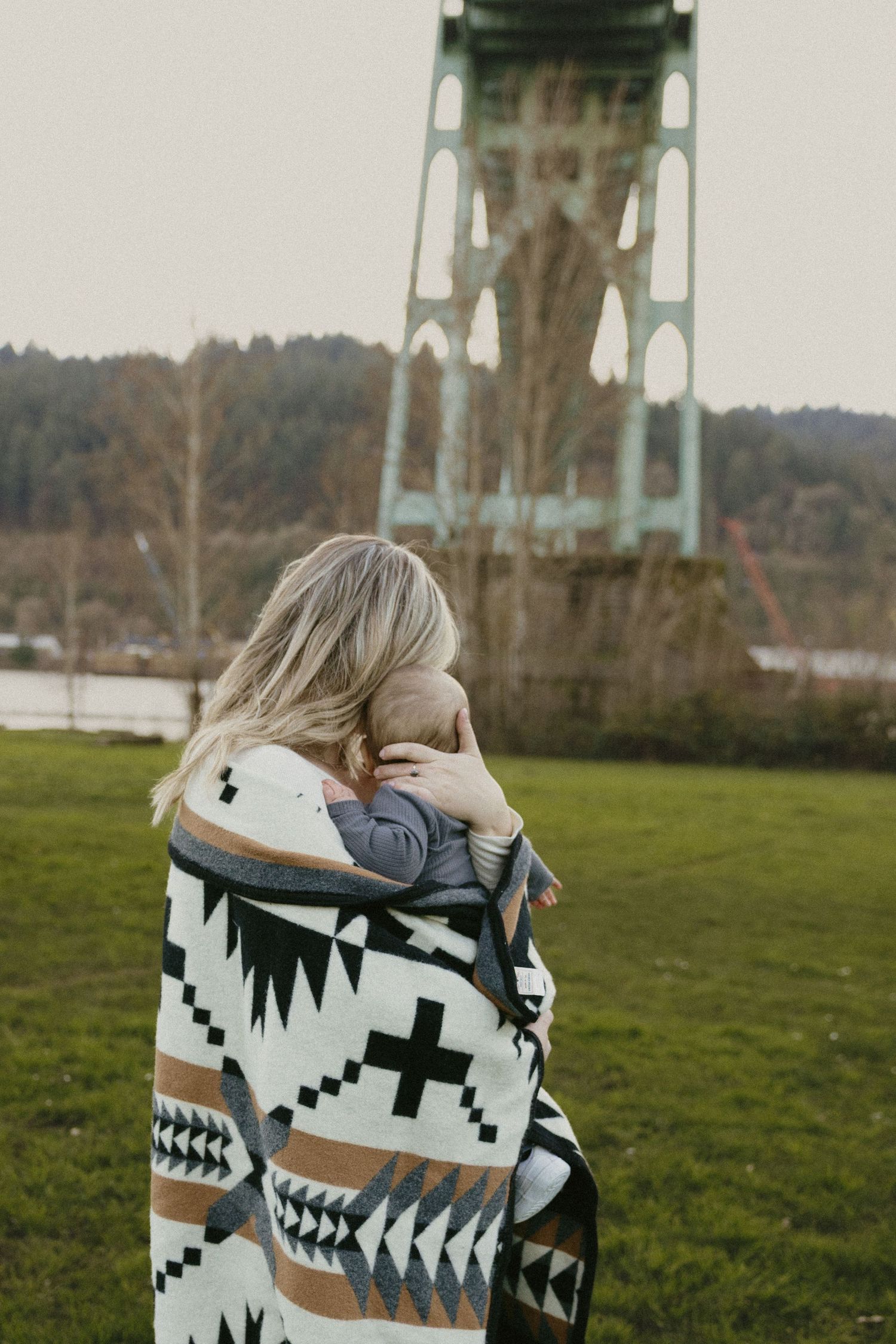 Mother wrapped in Native American pattern blanket cuddling baby facing St. Johns Bridge in Portland park.