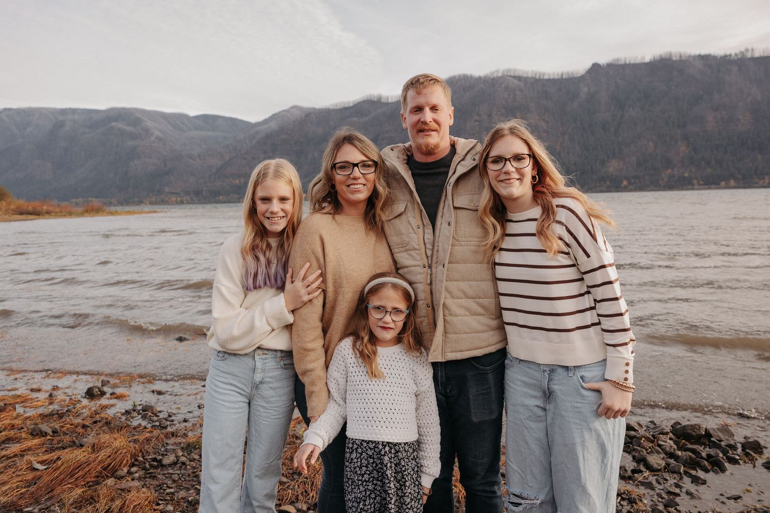 Smiling family of five stands together by a river with scenic mountain views behind them.