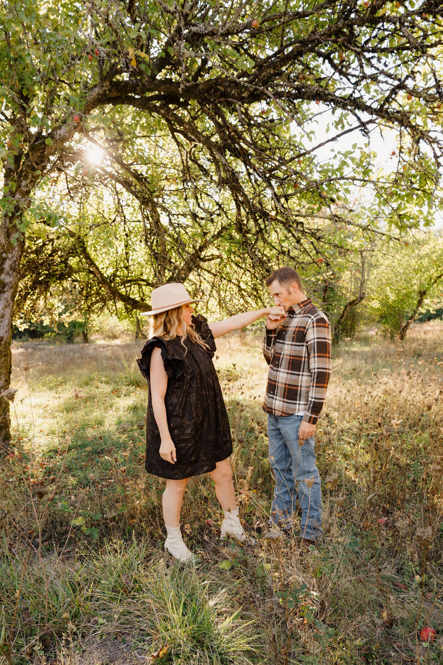 A couple dances playfully under a gnarled tree in a sunlit orchard field during golden hour.